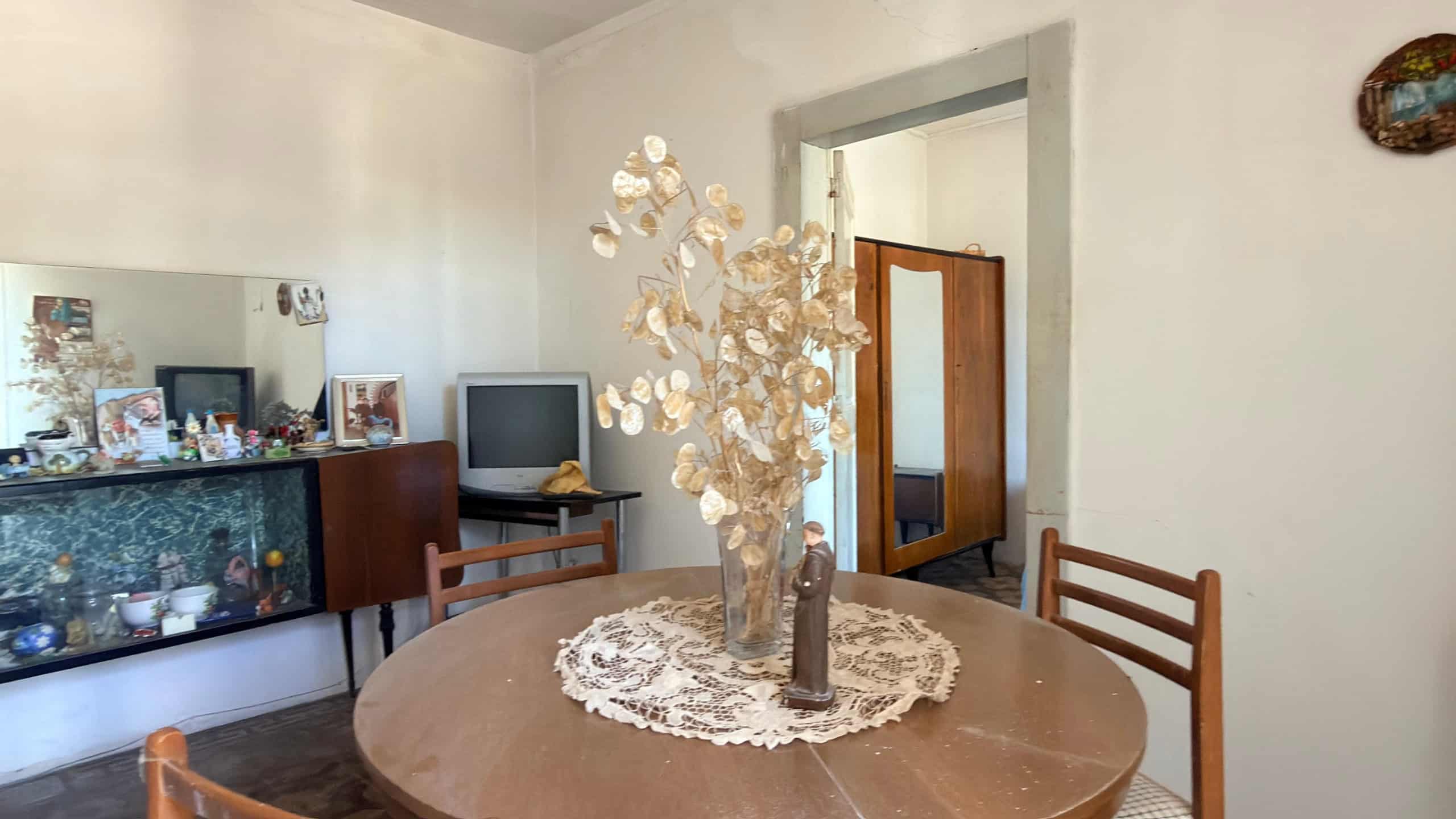 Tavolo rotondo in legno con decorazione floreale centrale e sedie in legno in soggiorno luminoso con pareti bianche e arredi classici.