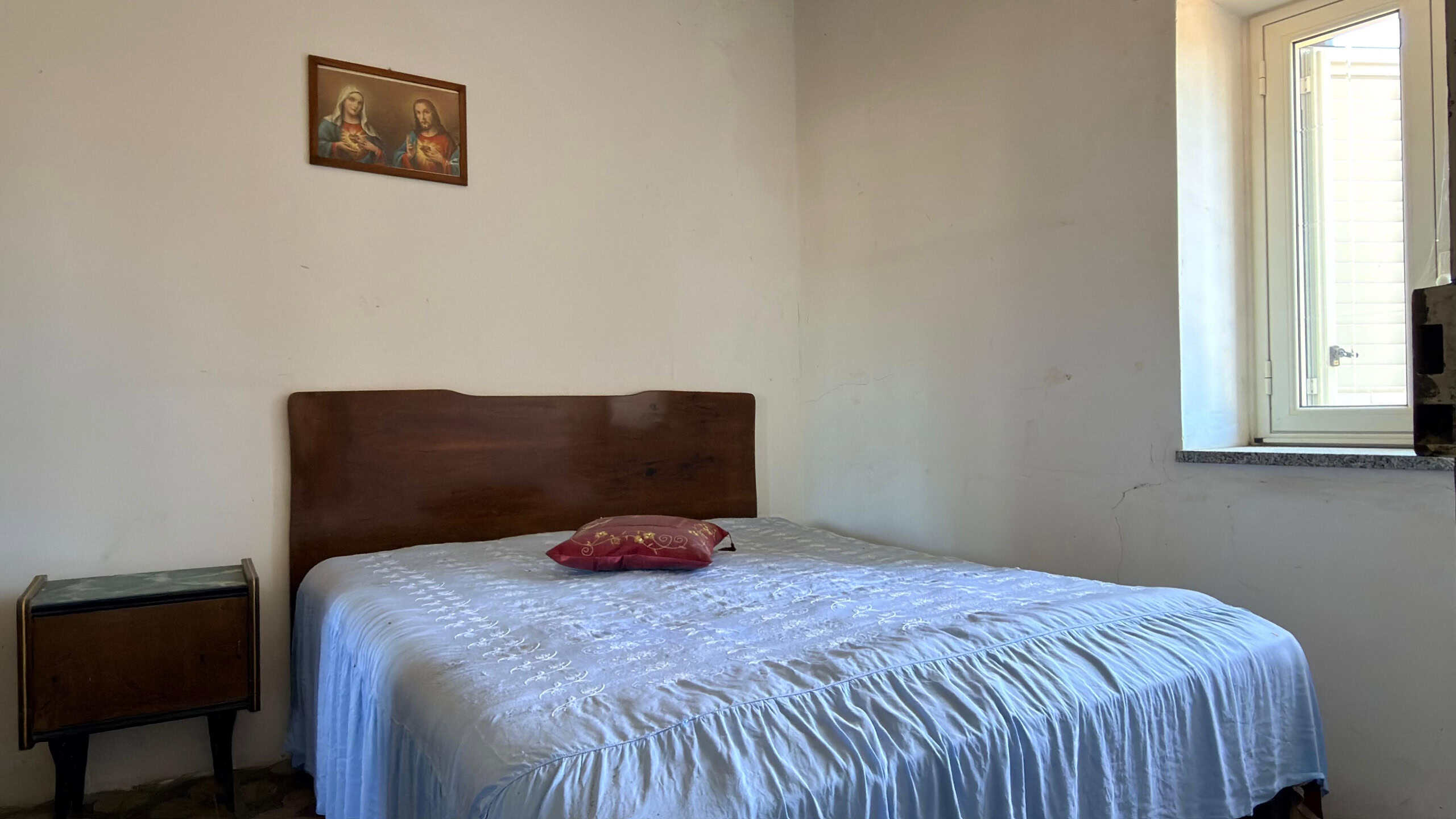 Il letto matrimoniale con testiera in legno in una stanza semplice e luminosa, con una finestra aperta e un quadro religioso appeso alla parete.