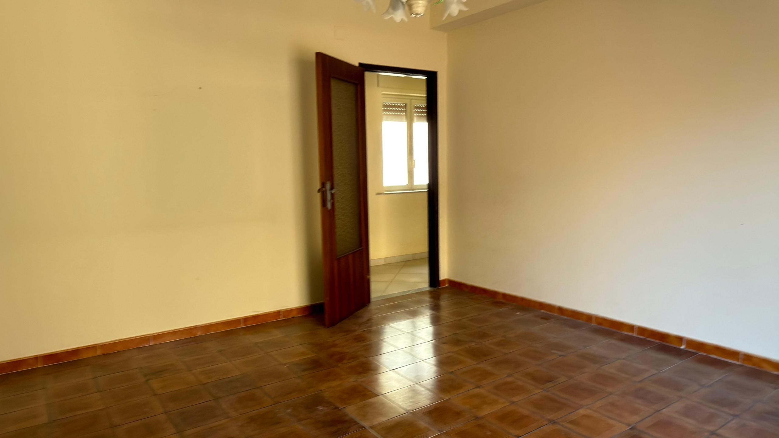 Ampia stanza vuota con pareti beige e pavimento in piastrelle di ceramica marrone, con porta in legno aperta che conduce a una stanza luminosa con finestra. Ideale per ristrutturazione o arredamento p.