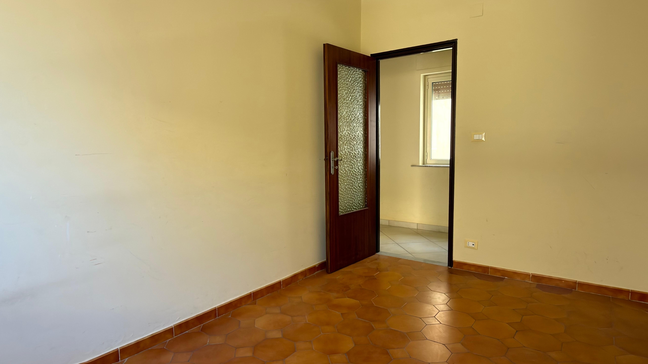 Ampia stanza vuota con pareti beige e pavimento in piastrelle di terracotta, porta in legno aperta che conduce a una stanza luminosa con finestra. Ideale per arredamento e ristrutturazione immobili.