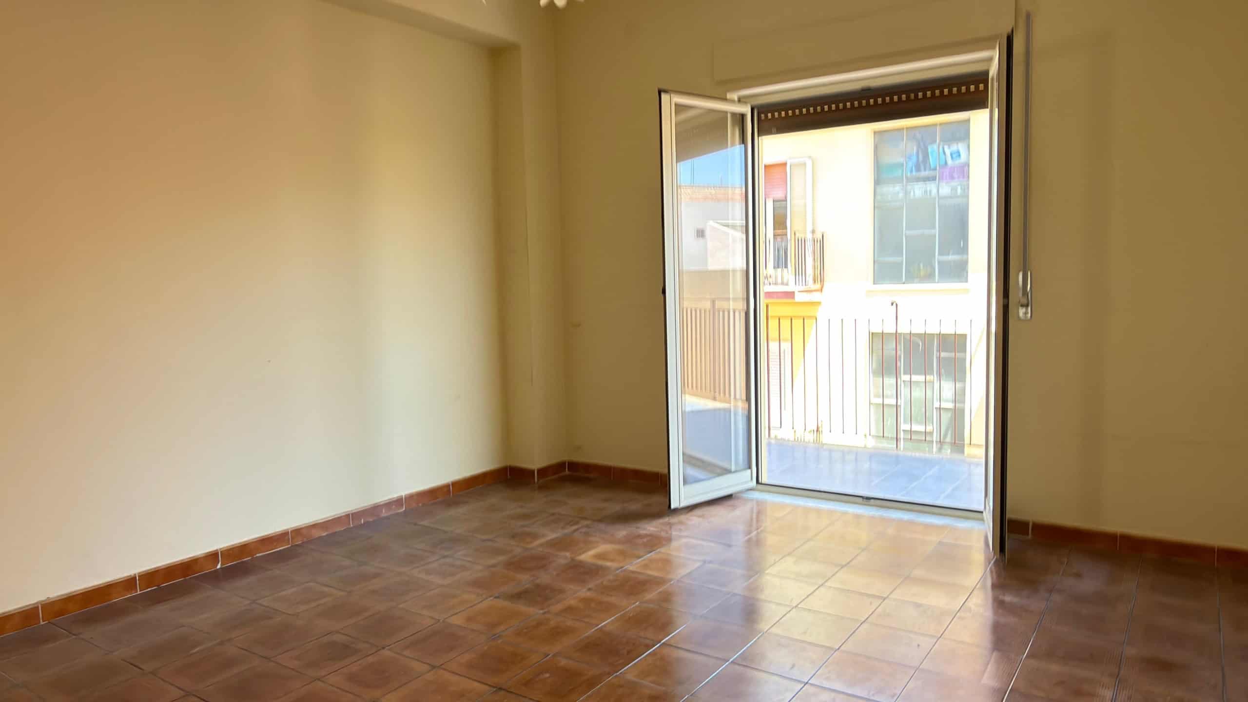 Ampio soggiorno vuoto con porta finestra che dà su un balcone, pavimento in piastrelle marroni e pareti beige, luce naturale che illumina l'ambiente.