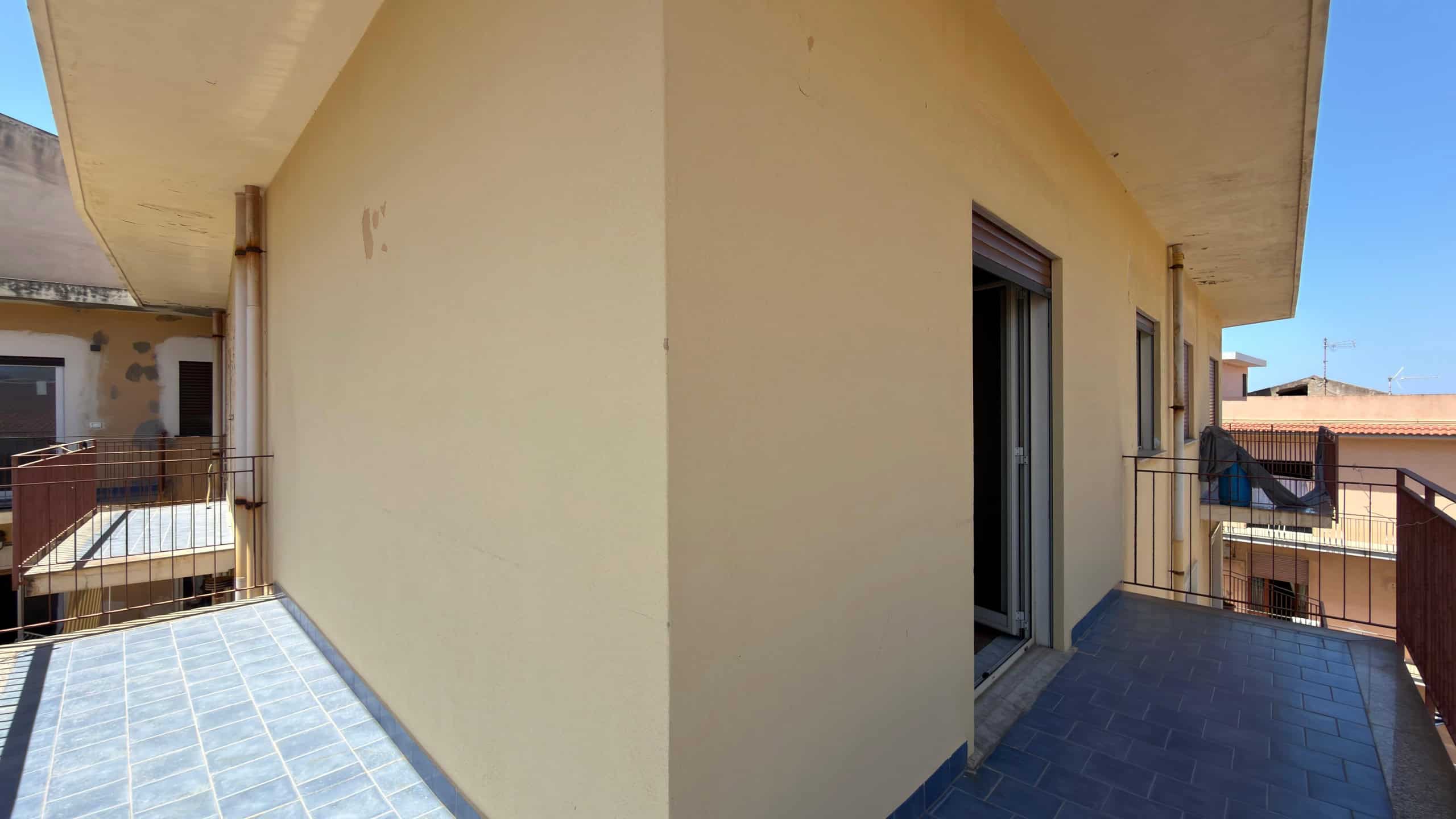 Balcone di un appartamento con vista sulla città, pavimentato in piastrelle scure e pareti beige, con accesso tramite porta finestra e ringhiera in metallo, in un contesto urbano soleggiato.