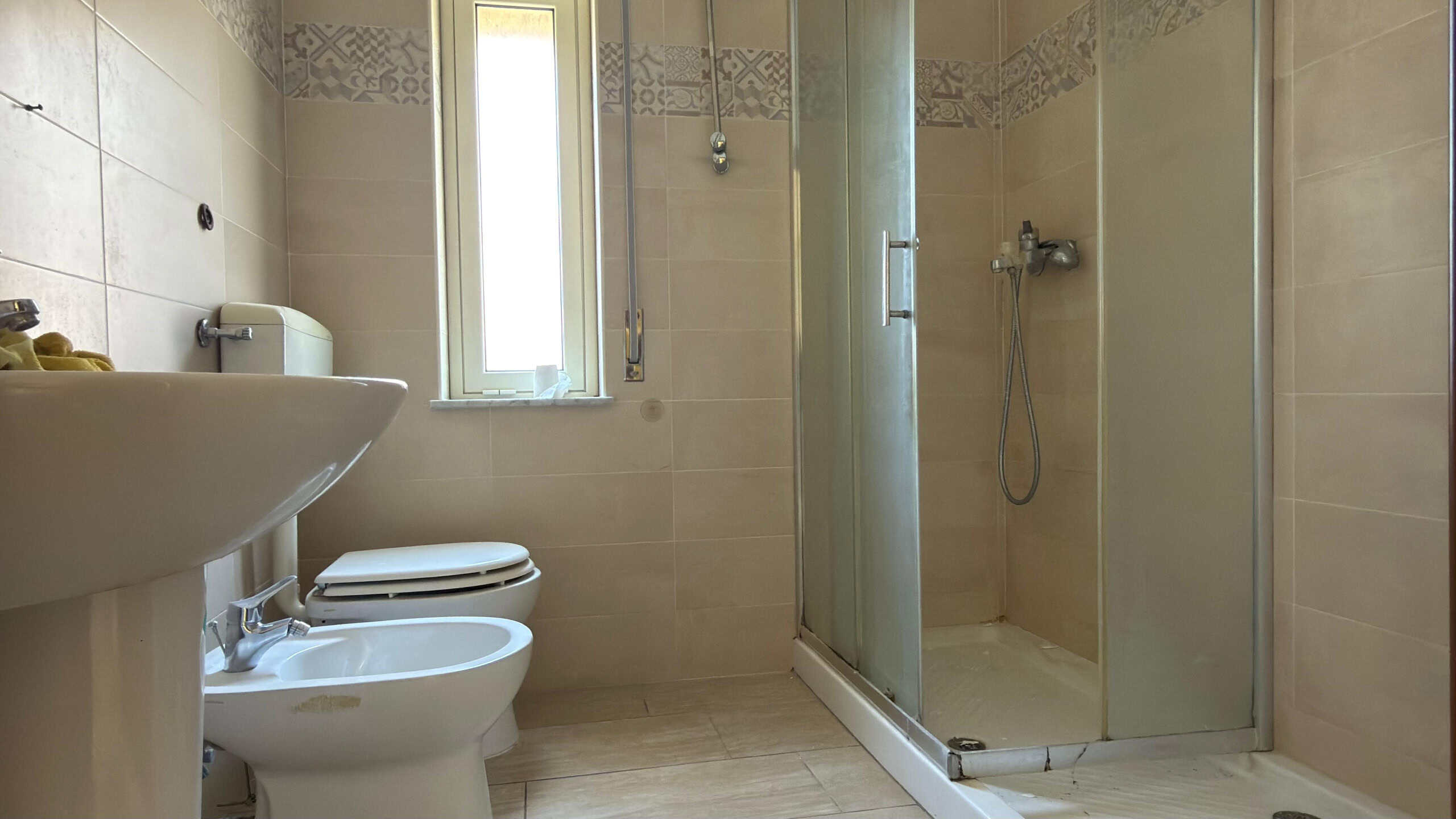 Il bagno moderno con doccia in vetro, lavabo e WC, illuminato dalla luce naturale proveniente dalla finestra, presenta piastrelle beige e uno stile pulito e funzionale.