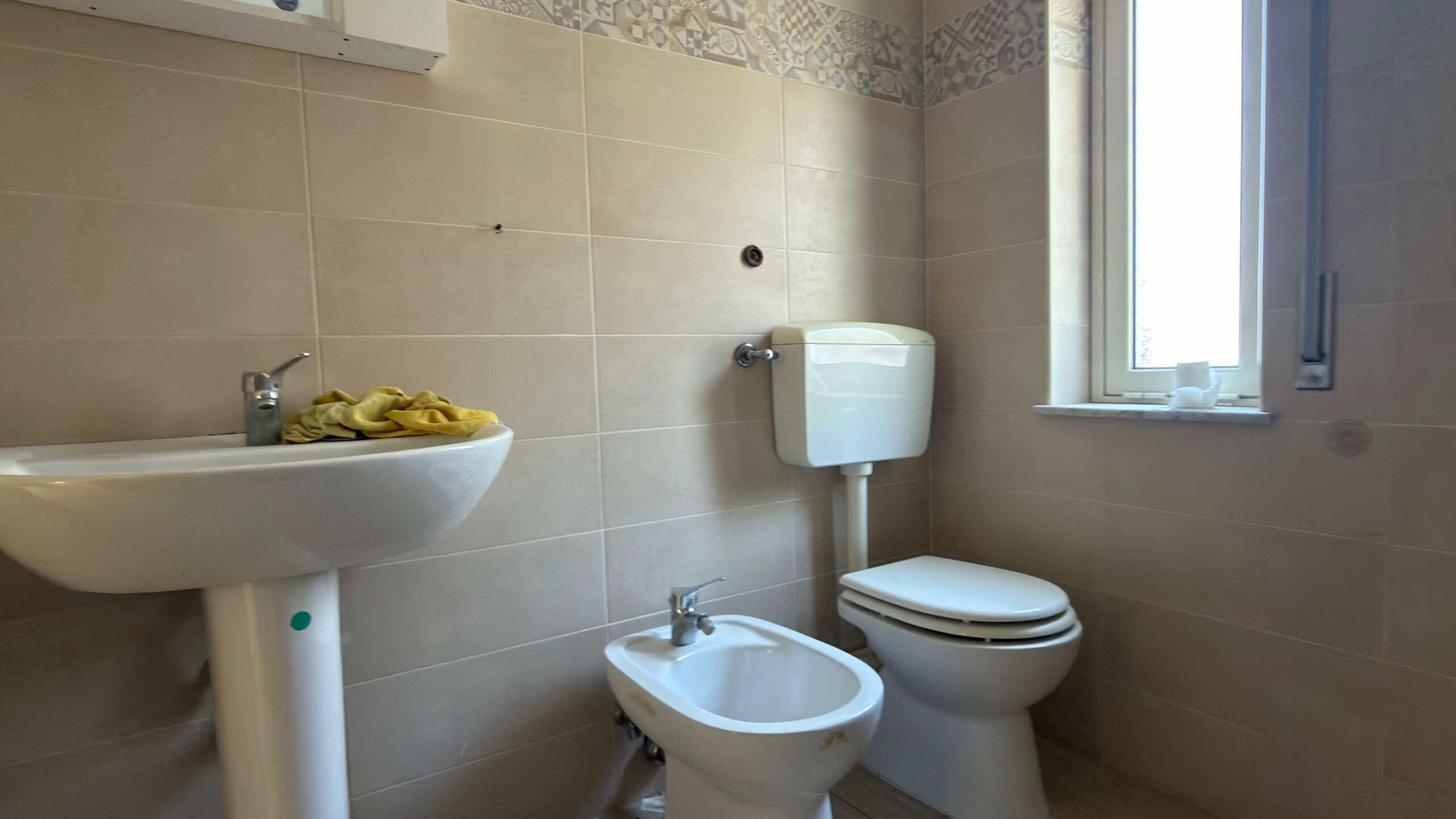 Lavabo e WC in un bagno moderno con pareti rivestite in piastrelle beige, finestra luminosa e ambiente pulito, ideale per ristrutturazioni e soluzioni abitative di qualità.