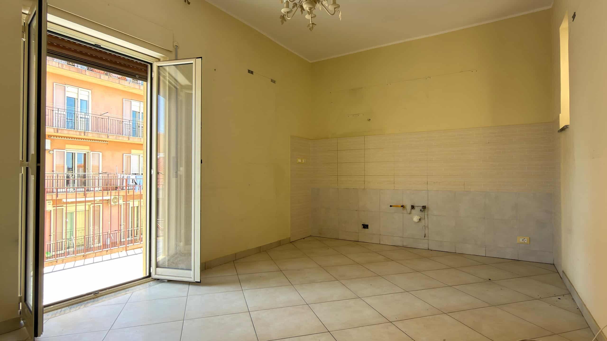 Ampia stanza luminosa con porta finestra che dà su un balcone, pareti chiare e pavimento in piastrelle, ideale per ristrutturazione e arredamento moderno.