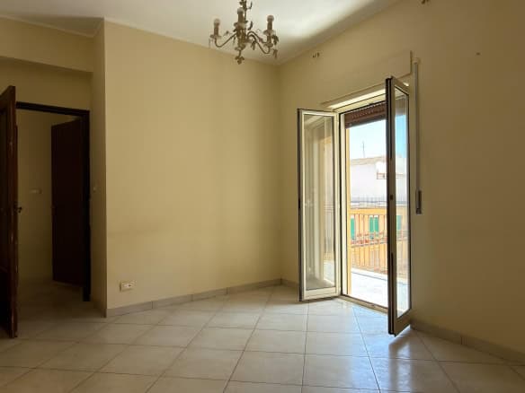Ampia stanza luminosa con porta finestra che dà su un balcone, pareti beige e pavimento in piastrelle chiaro, ideale per la vendita o l'affitto di immobili residenziali.