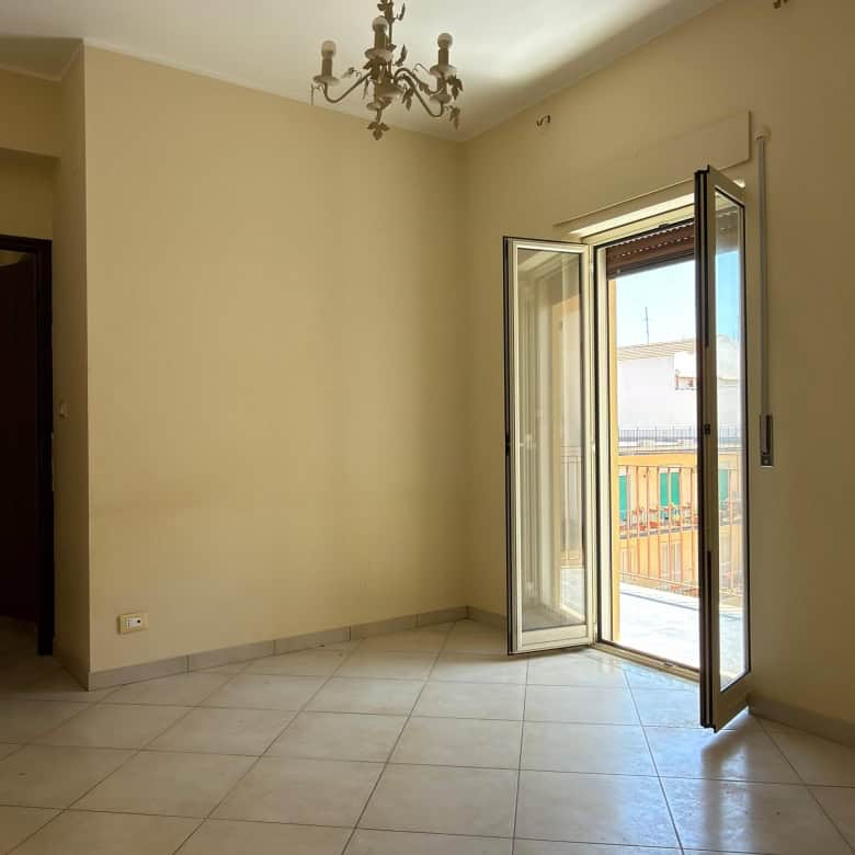 Ampia stanza luminosa con porta finestra che dà su un balcone, pareti beige e pavimento in piastrelle chiaro, ideale per la vendita o l'affitto di immobili residenziali.