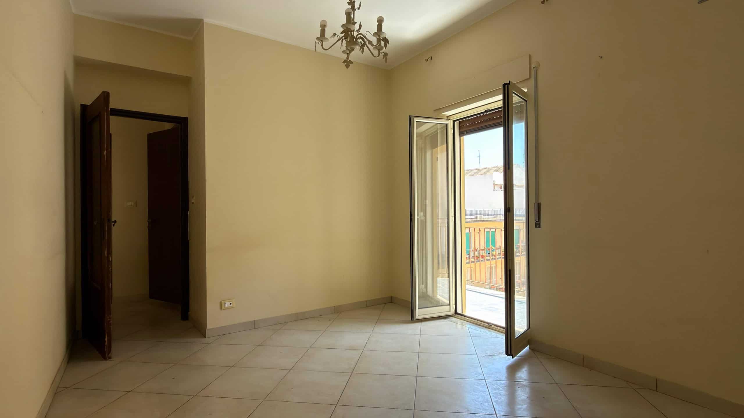 Ampia stanza luminosa con porta finestra che dà su un balcone, pareti beige e pavimento in piastrelle chiaro, ideale per la vendita o l'affitto di immobili residenziali.