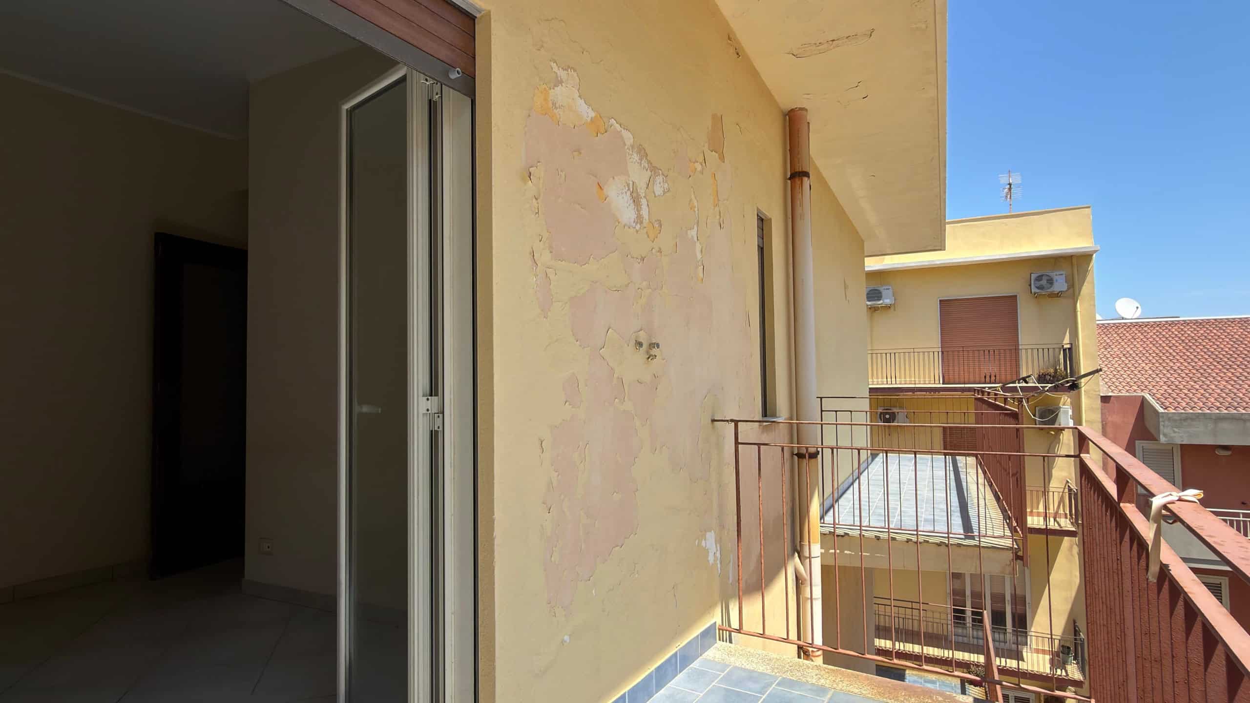 Balcone di un appartamento con pareti esterne in condizioni di deterioramento, vista su edifici circostanti e cielo sereno, in un contesto urbano.