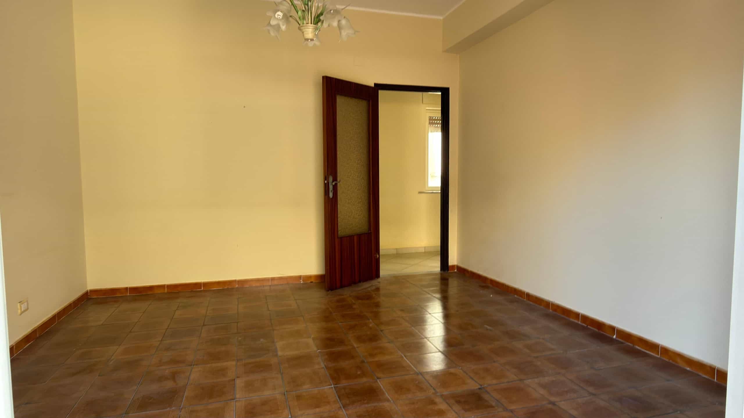 Spazio vuoto in una stanza con pareti beige, pavimento in piastrelle marroni e porta in legno aperta, pronta per arredamento e personalizzazione.