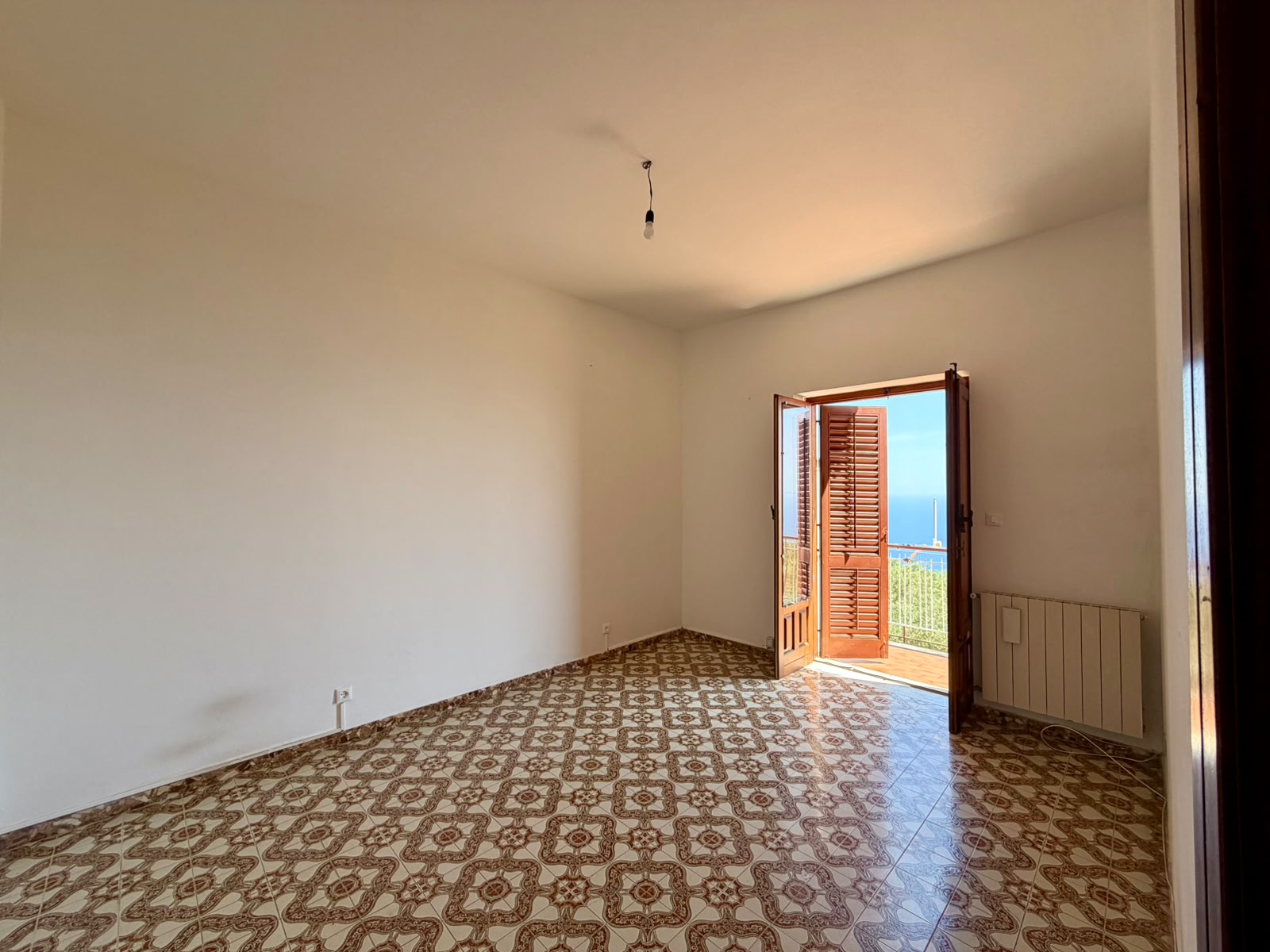 Stanza vuota con pareti bianche, pavimento in piastrelle decorato e porta finestra che si apre su un balcone con vista panoramica.