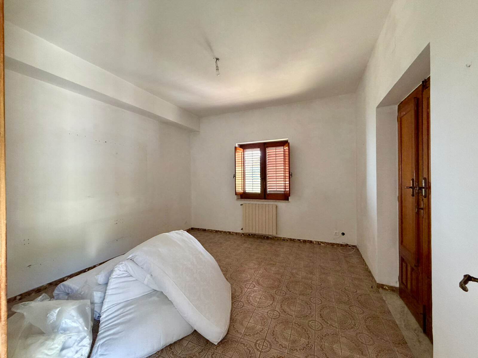 Ampio soggiorno vuoto con pareti bianche, finestra con persiane in legno e porta in legno, pavimento in piastrelle decorative, ambiente luminoso e spazioso.