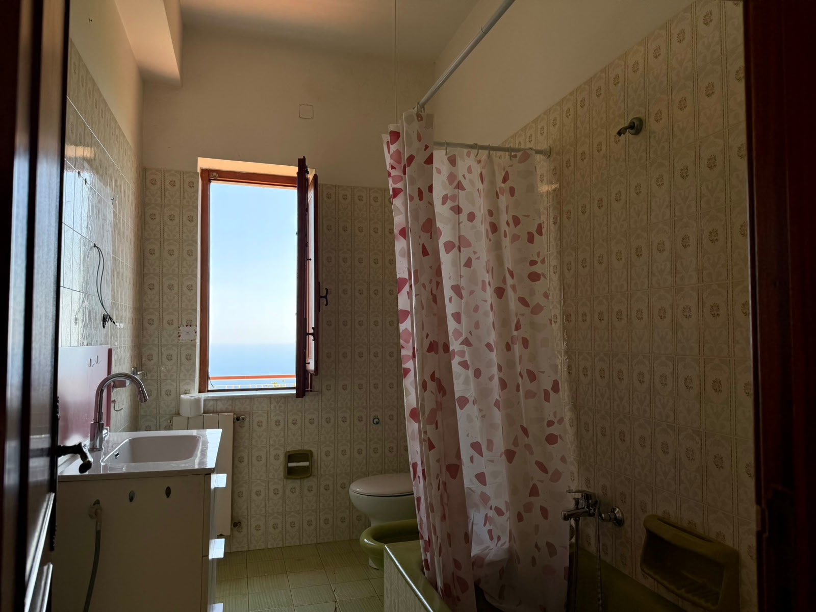 Doccia e lavabo in bagno con finestra aperta, luce naturale e pareti rivestite con piastrelle vintage.