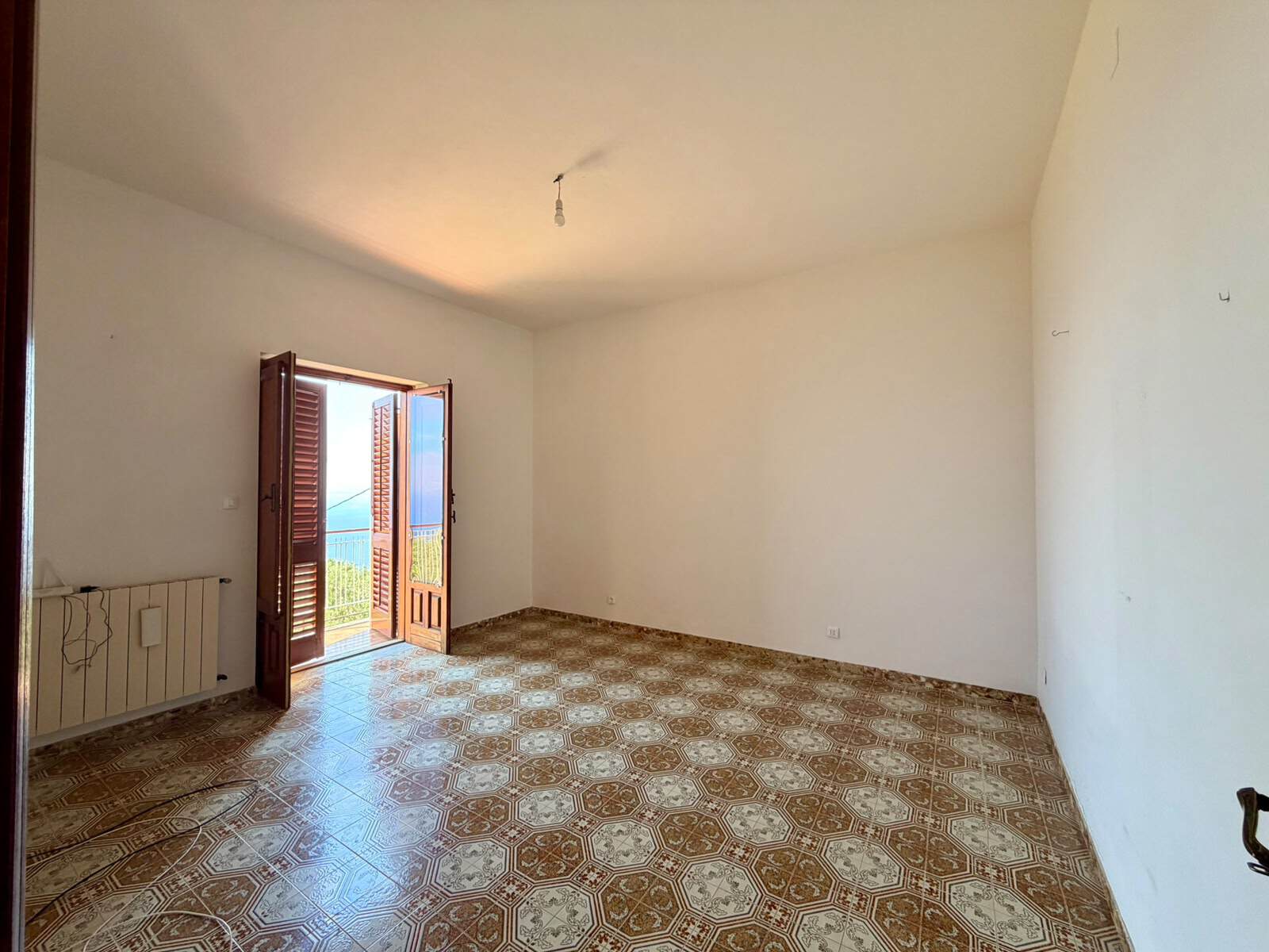 Stanza vuota con porta finestra che apre su un balcone con vista mare, pavimento in piastrelle decorato, pareti bianche e soffitto semplice.