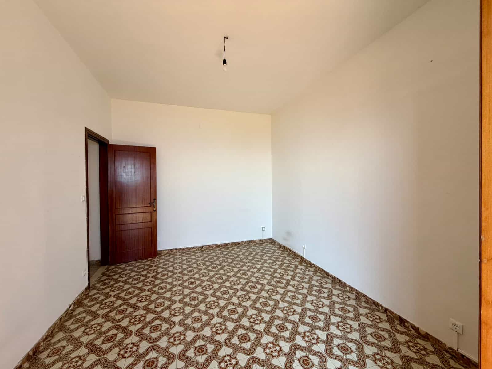 Appartamento vuoto con pareti bianche e pavimento in piastrelle decorative, porta in legno scuro, stanza luminosa e spaziosa, ideale per ristrutturazione o arredamento personalizzato.