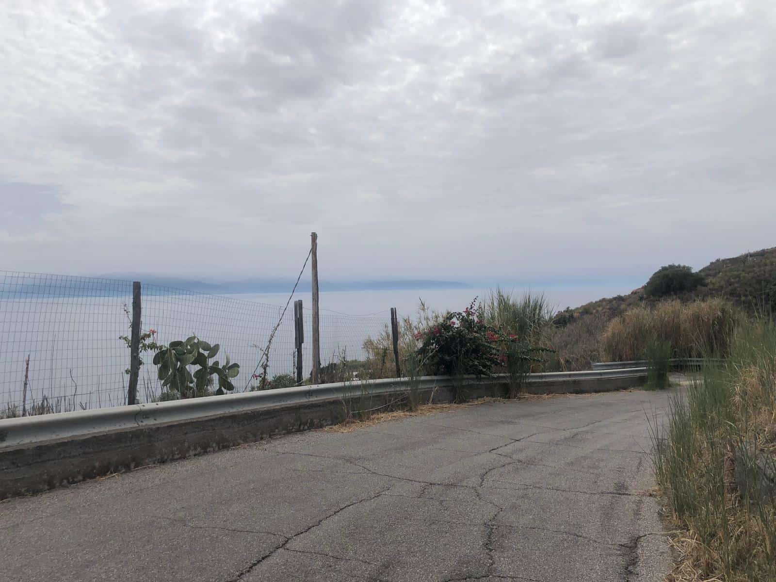 Barriera stradale e vista panoramica sul mare con cielo nuvoloso, natura e paesaggio costiero in background, strada asfaltata in primo piano, ambiente tranquillo e suggestivo, ideale per case al mare.