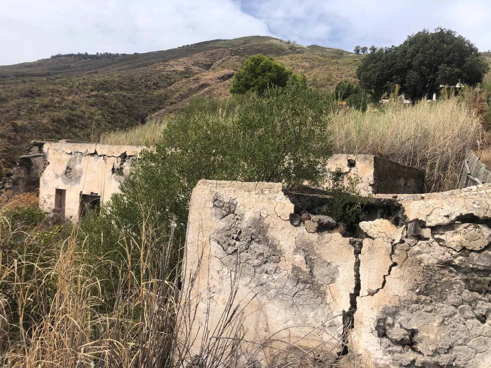 Rovine di un edificio abbandonato in un paesaggio collinare con vegetazione e montagne sullo sfondo.