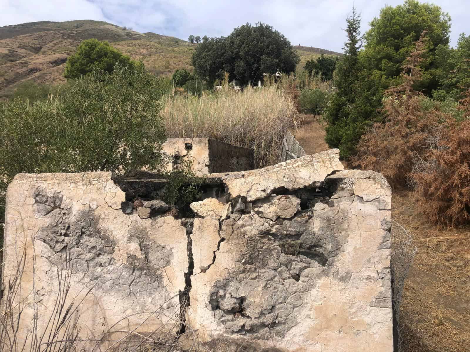 Viti in pietra e rovine di un'antica struttura rurale immersa in un paesaggio collinare con vegetazione mediterranea, alberi e arbusti, sotto un cielo nuvoloso.