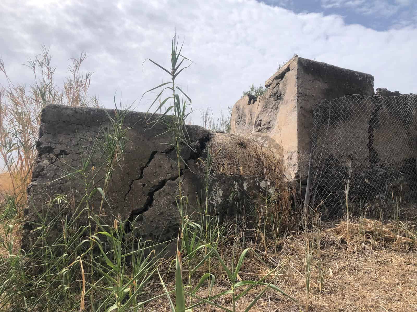 Vecchio edificio in rovina con muri di cemento e recinzione in rete metallica, circondato da erba secca e piante selvatiche, sotto un cielo nuvoloso, rappresenta un sito abbandonato o storico.