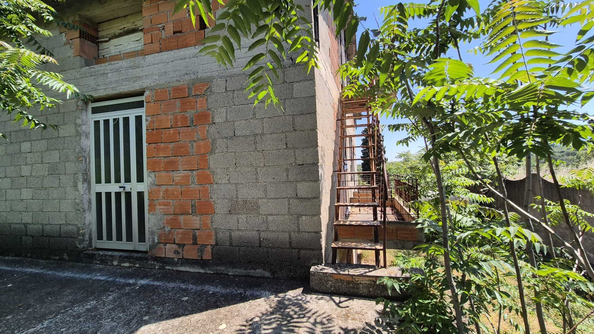 Ampia porta bianca in metallo con grate, ingresso di una casa in costruzione, ambiente esterno con scale in ferro e vegetazione rigogliosa, immagine di un edificio in fase di completamento.