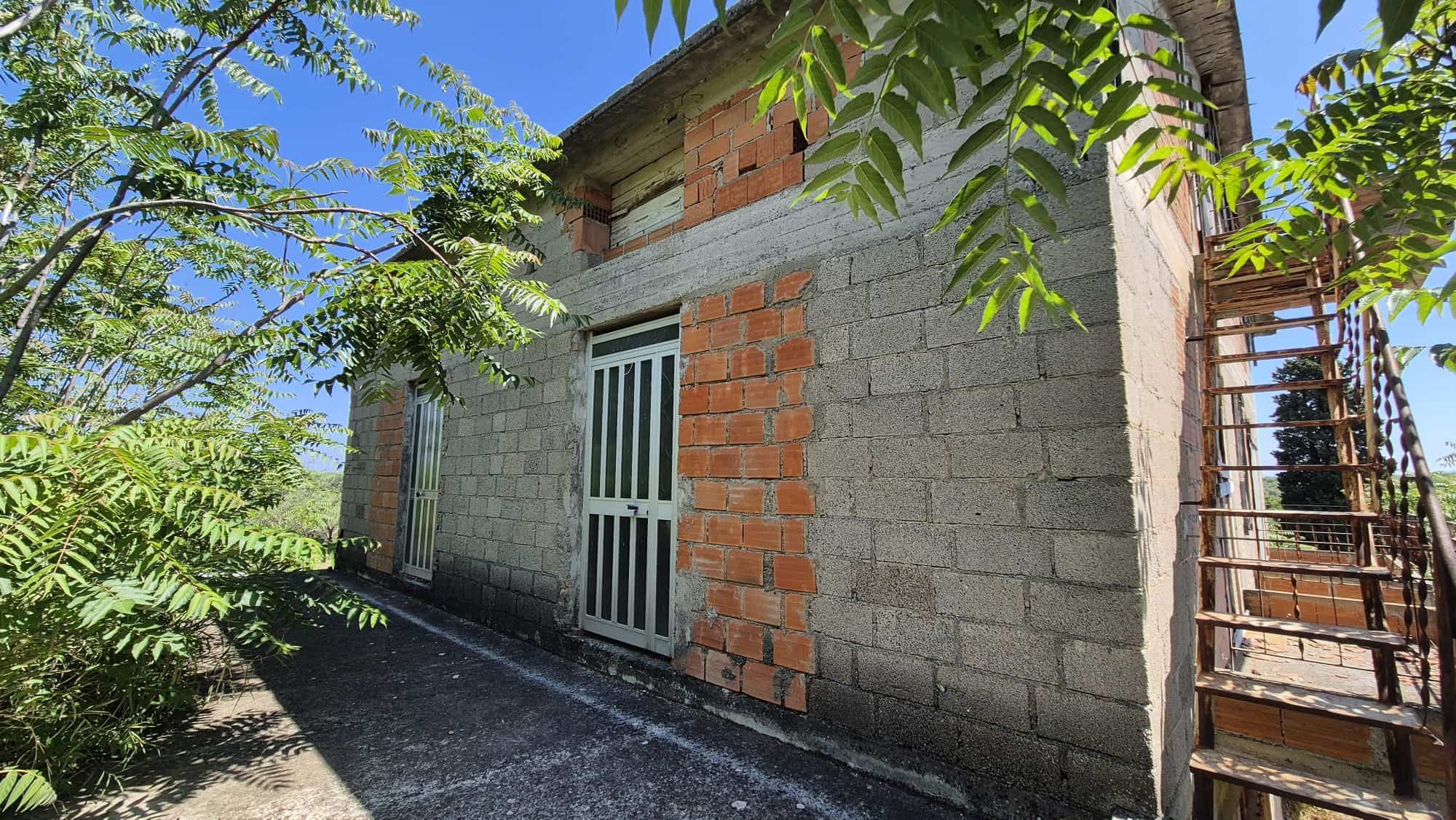 Ampia casa in costruzione con pareti in mattoni e cemento, circondata da vegetazione verde, con scala esterna in ferro e accesso principale con porta bianca.