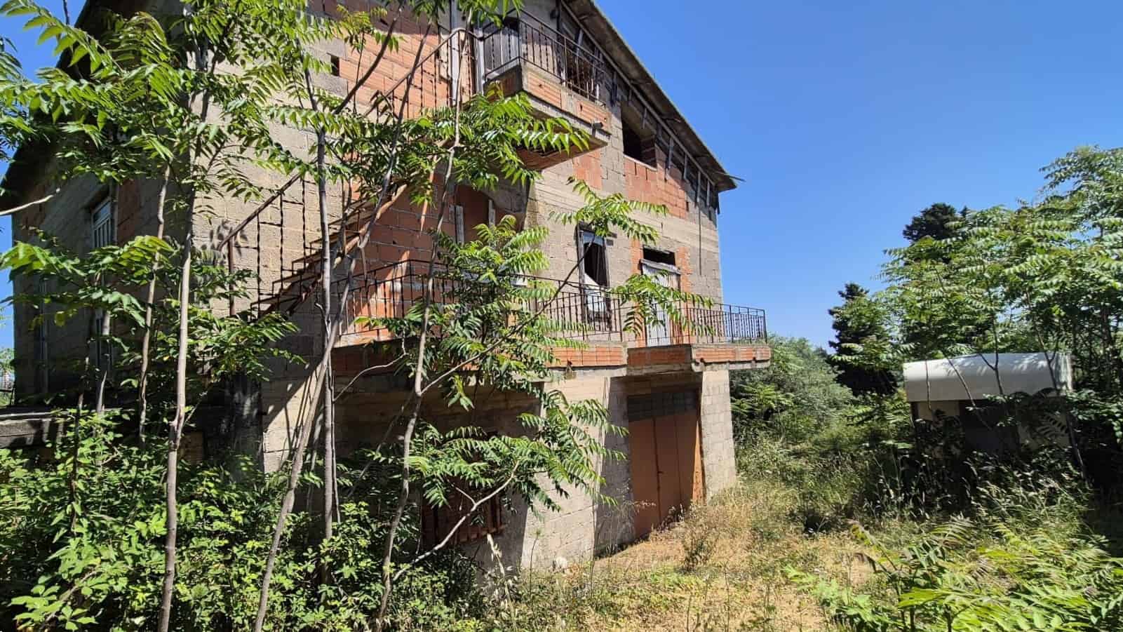 Ampia casa in costruzione con balconi e terrazze, circondata da vegetazione verde e alberi, sotto un cielo azzurro, pronta per essere ristrutturata o completata.