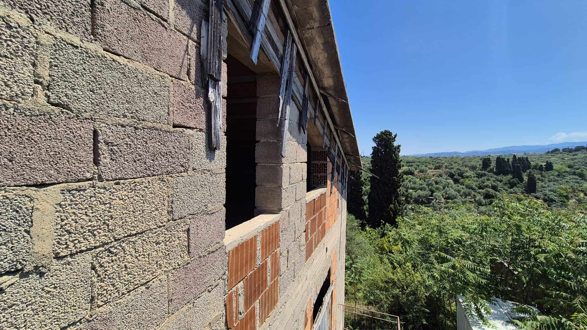 Scorcio di un edificio in costruzione con pareti in mattoni e blocchi di cemento, circondato da un paesaggio naturale verde e un cielo sereno, rappresentando un progetto edilizio in fase di sviluppo.