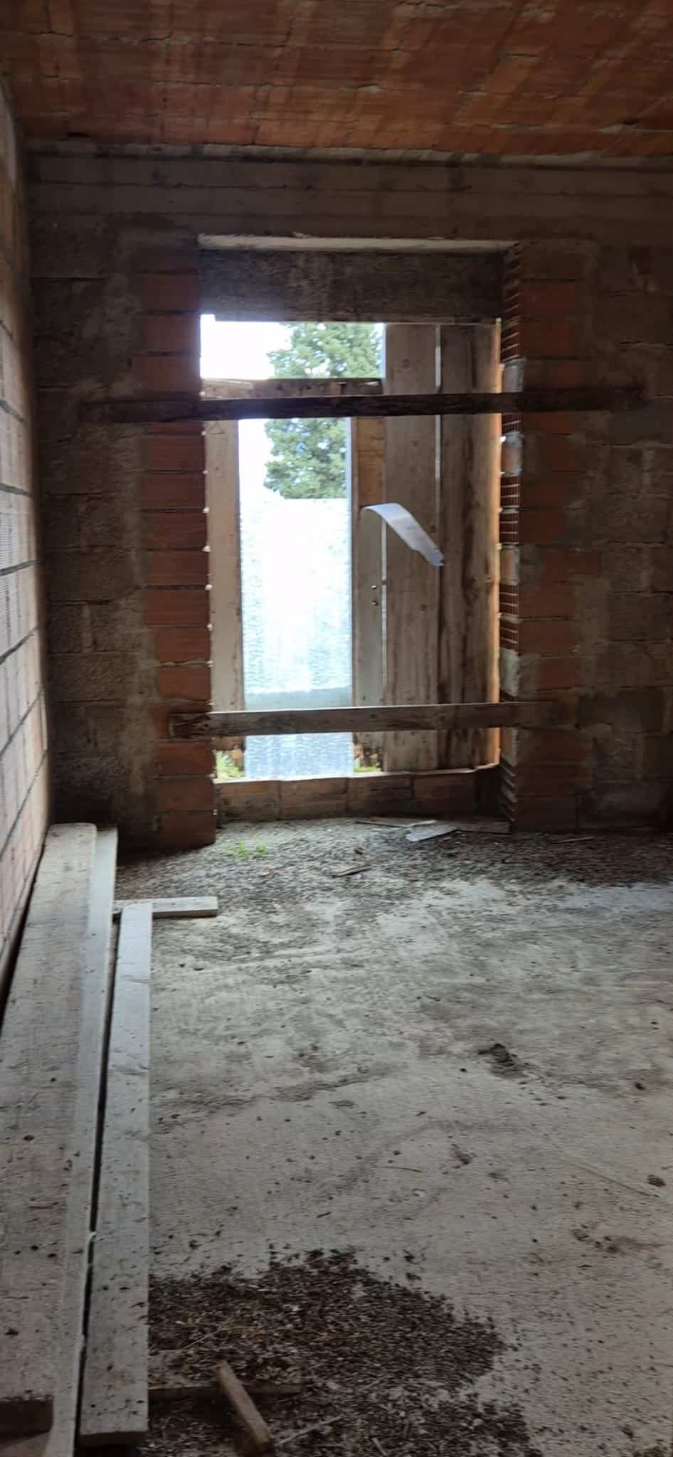 Prospettiva di una finestra in costruzione con struttura in mattoni e cemento, all’interno di un edificio in fase di ristrutturazione, con vista su un albero e il cielo.