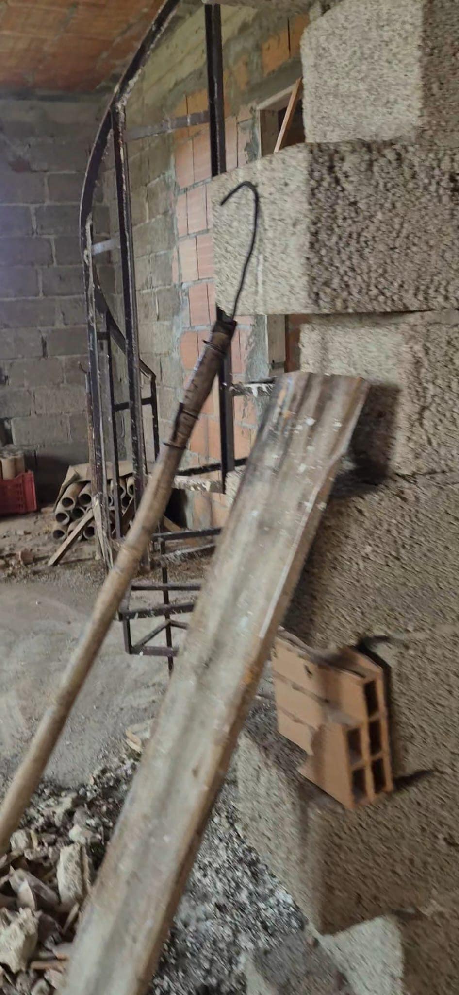 Palo di legno appoggiato contro un muro in costruzione, con attrezzi e materiali da cantiere sullo sfondo, in un ambiente di lavoro edilizio.