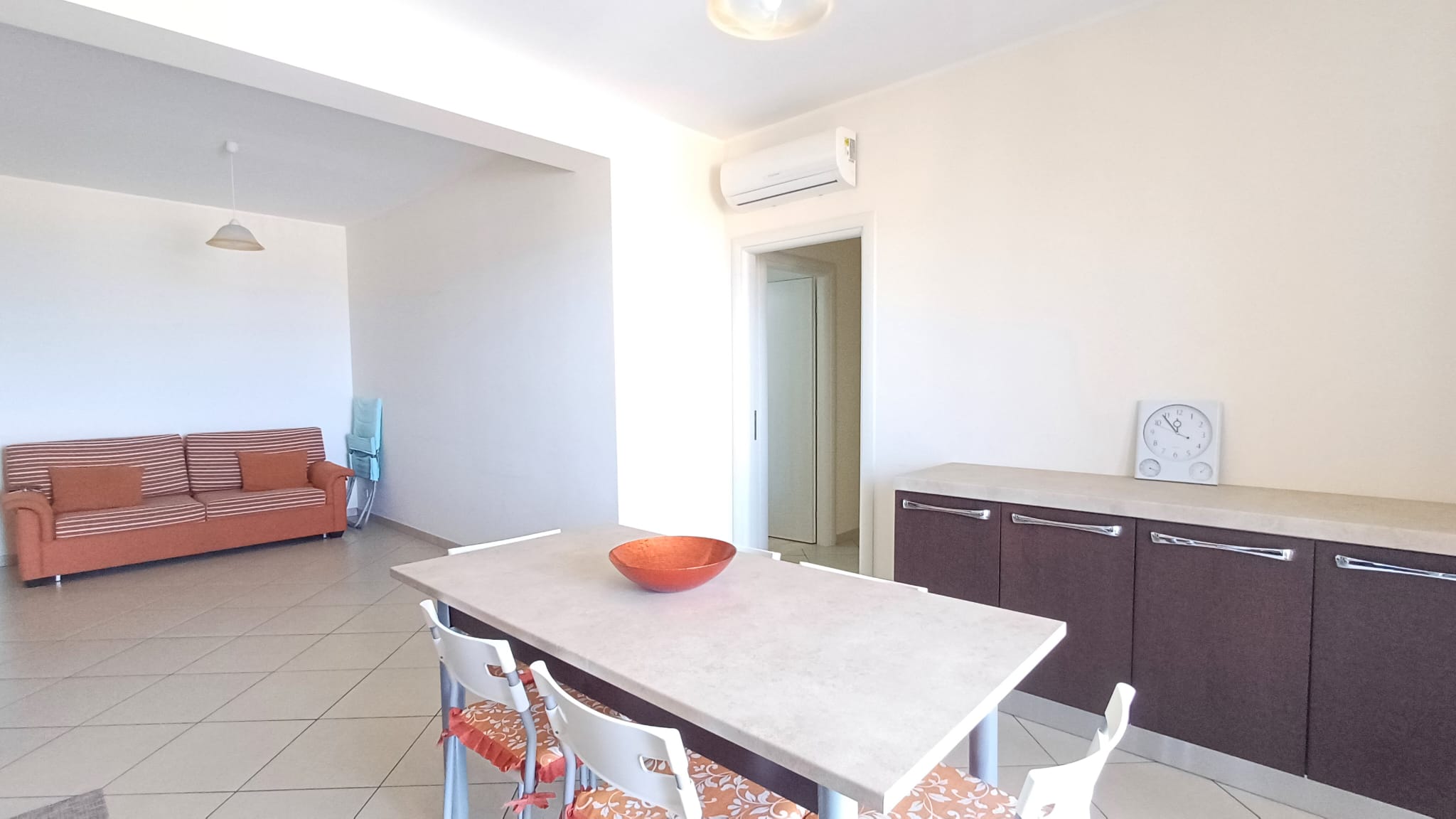 Ampio soggiorno moderno con divano arancione, tavolo centrale e cucina a vista, ambiente luminoso e accogliente, ideale per vivere e ricevere ospiti in stile contemporaneo.