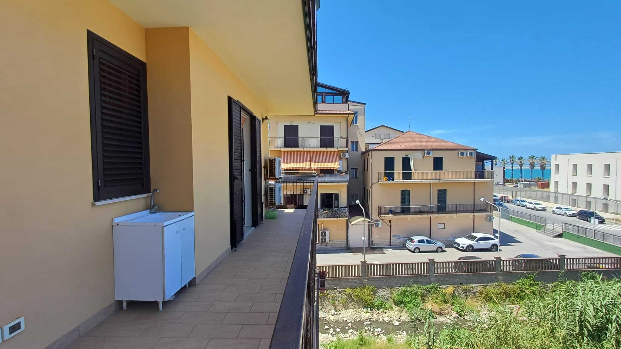 Spiaggia e mare visibili da un balcone di un appartamento in zona residenziale, con edifici moderni e parcheggio nelle vicinanze, in una giornata soleggiata e luminosa.