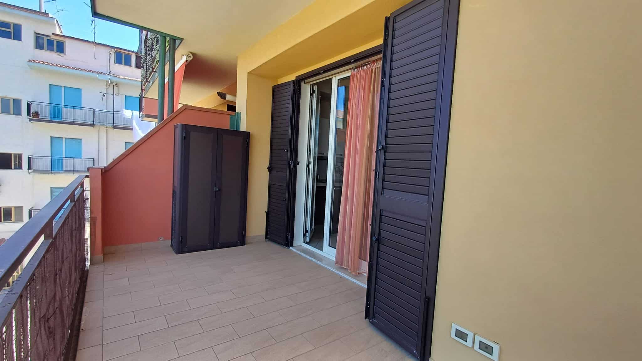 Ampio balcone con porte scorrevoli in vetro e persiane nere, affacciato su un complesso residenziale moderno, con pavimentazione in piastrelle e pareti gialle.
