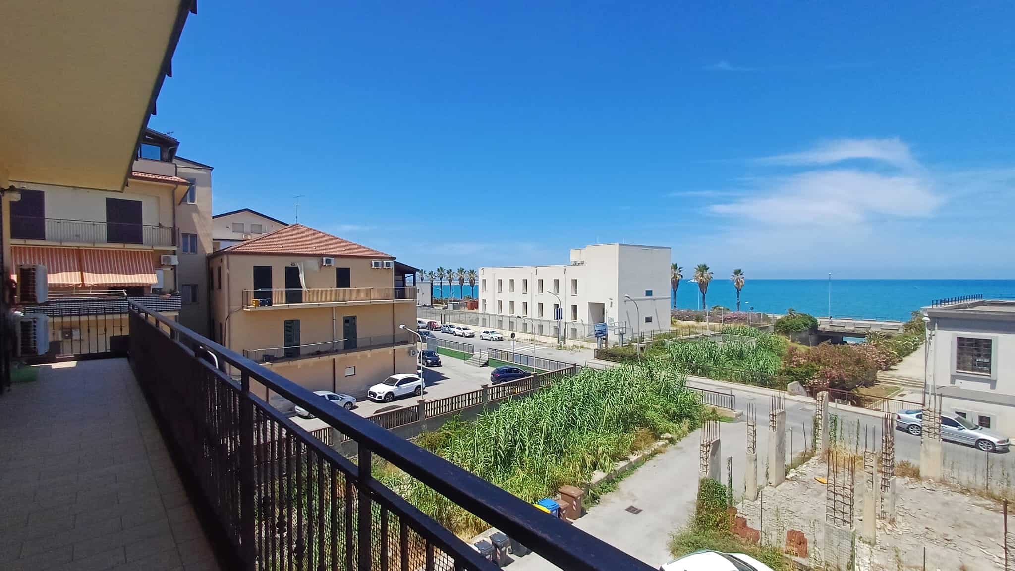 Vista panoramica di un appartamento con balcone che si affaccia sul mare, con edifici residenziali e spiaggia in lontananza, cielo sereno e atmosfera tranquilla.