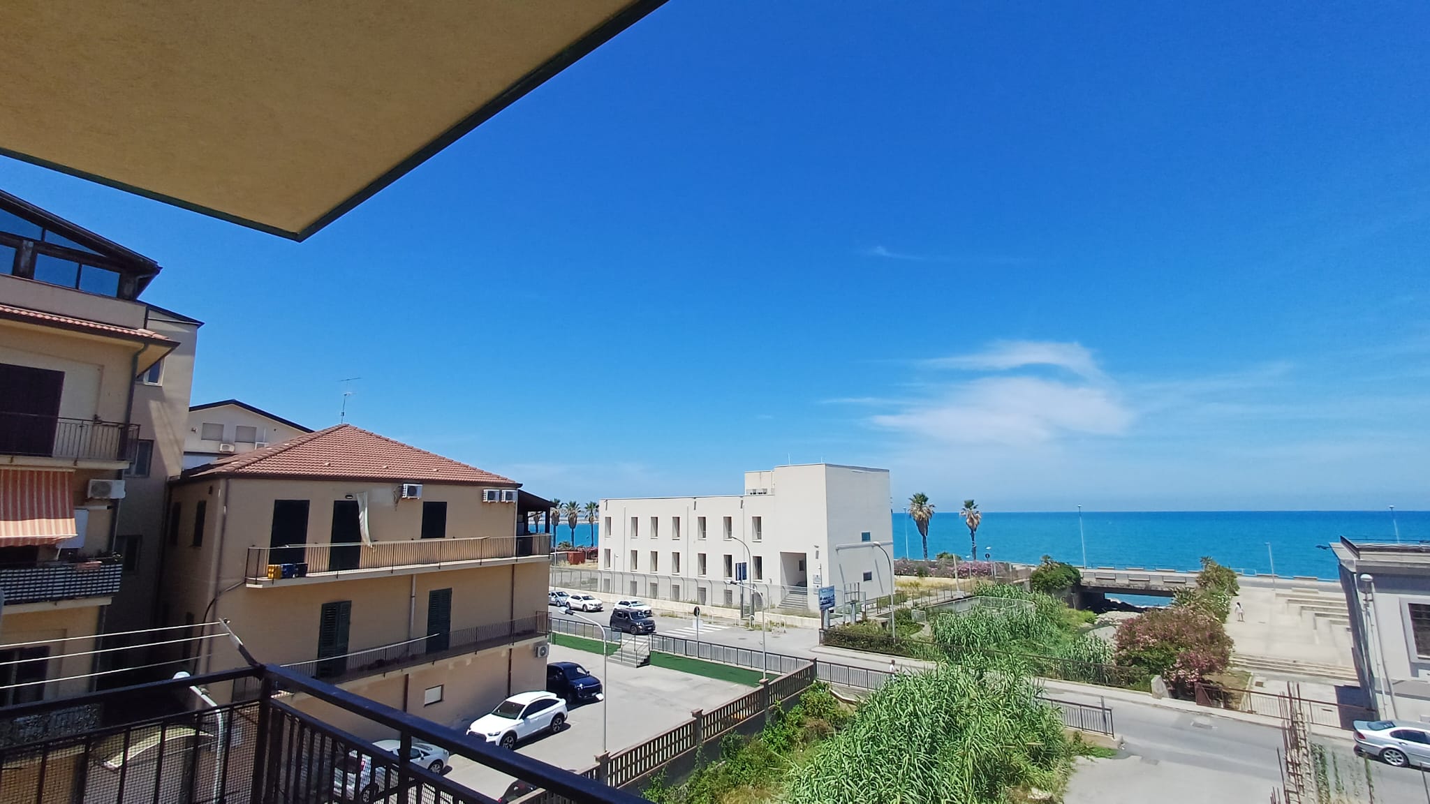 Spiaggia e mare visibili da un balcone di un appartamento a NextCasa, con edifici residenziali e un lungomare in lontananza, cielo sereno e atmosfera estiva.