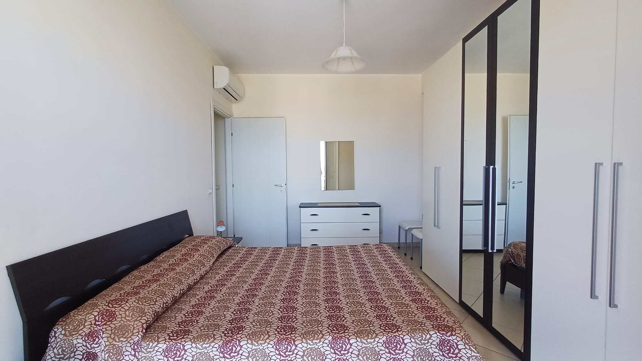 Letto matrimoniale in camera moderna con armadio a specchio e arredi minimalisti, ambientazione luminosa e accogliente, ideale per residenze di comfort e stile.