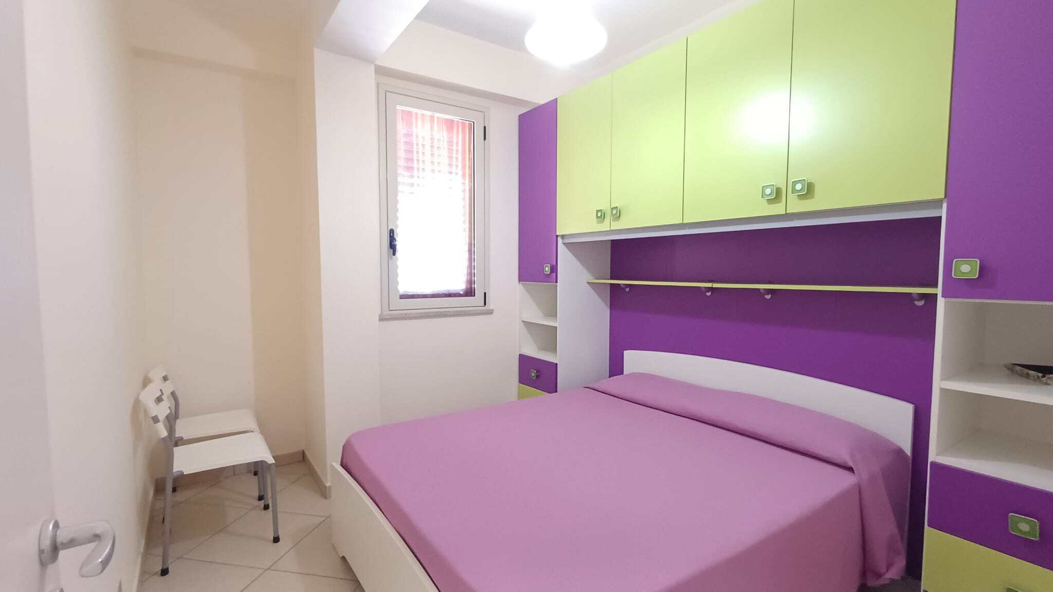 Ampio armadio a muro con ante color lime e viola in camera da letto moderna, con letto matrimoniale e finestra luminosa.