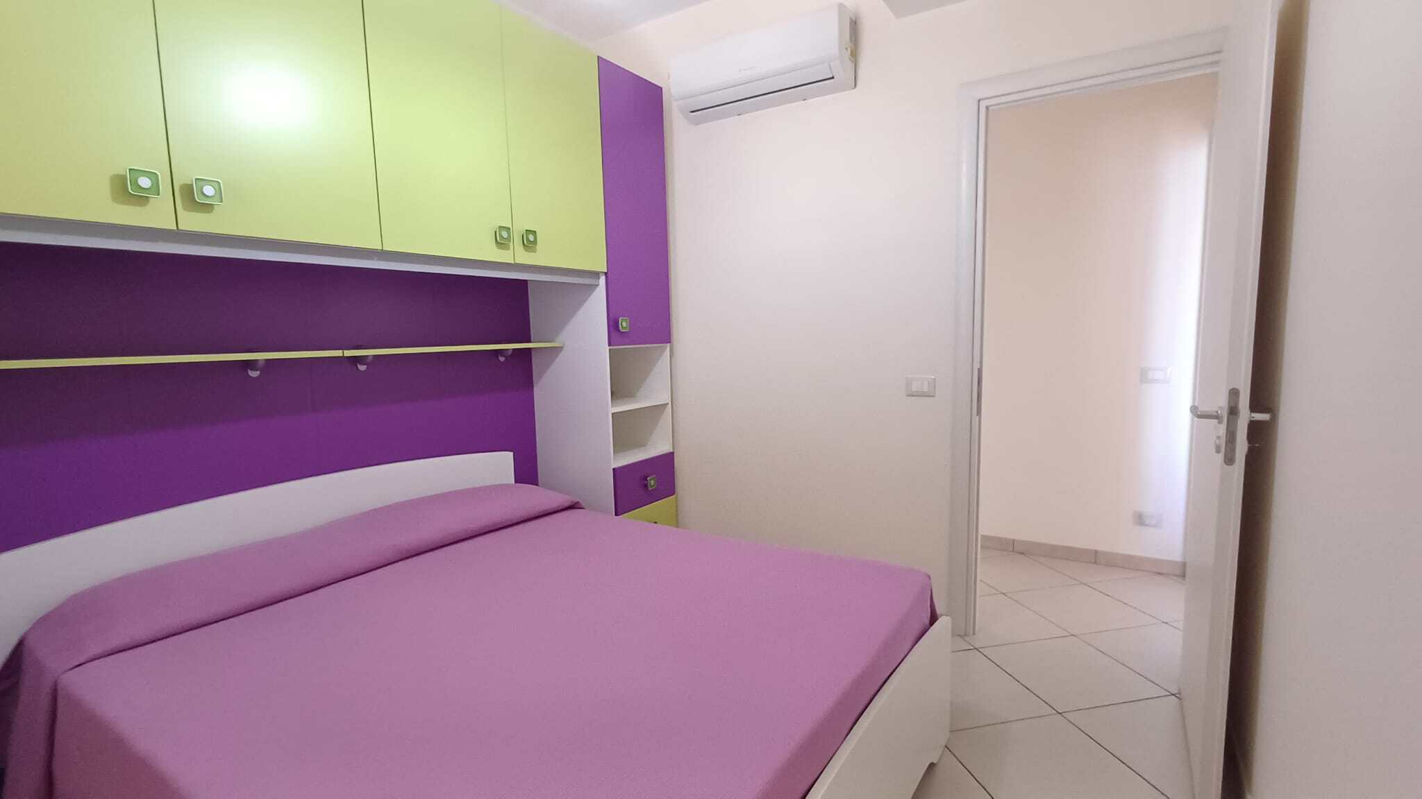 L'immagine mostra una camera da letto moderna con pareti colorate in verde e viola, un letto con biancheria viola, armadi a muro e aria condizionata. Spazio luminoso e accogliente, ideale per soggiorn.