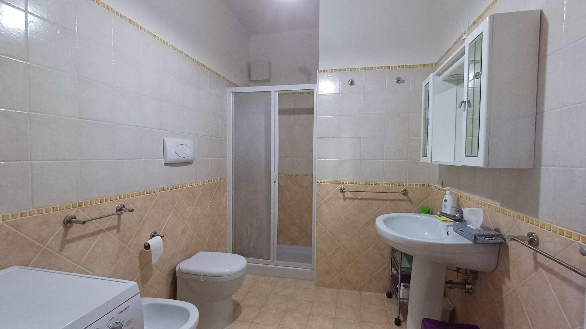 Lavabo e servizi igienici in un bagno moderno con pareti beige e accessori funzionali, inclusa una doccia con porta in vetro e un mobiletto con specchio sopra il lavandino.