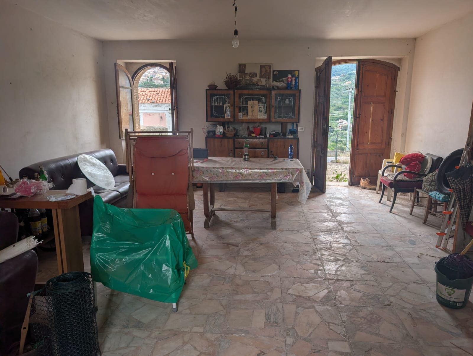 Il soggiorno di una casa rustica con pareti bianche, pavimento in pietra, finestre e porta in legno, arredato con divani, sedie e mobili vintage, con vista su un paesaggio collinare.