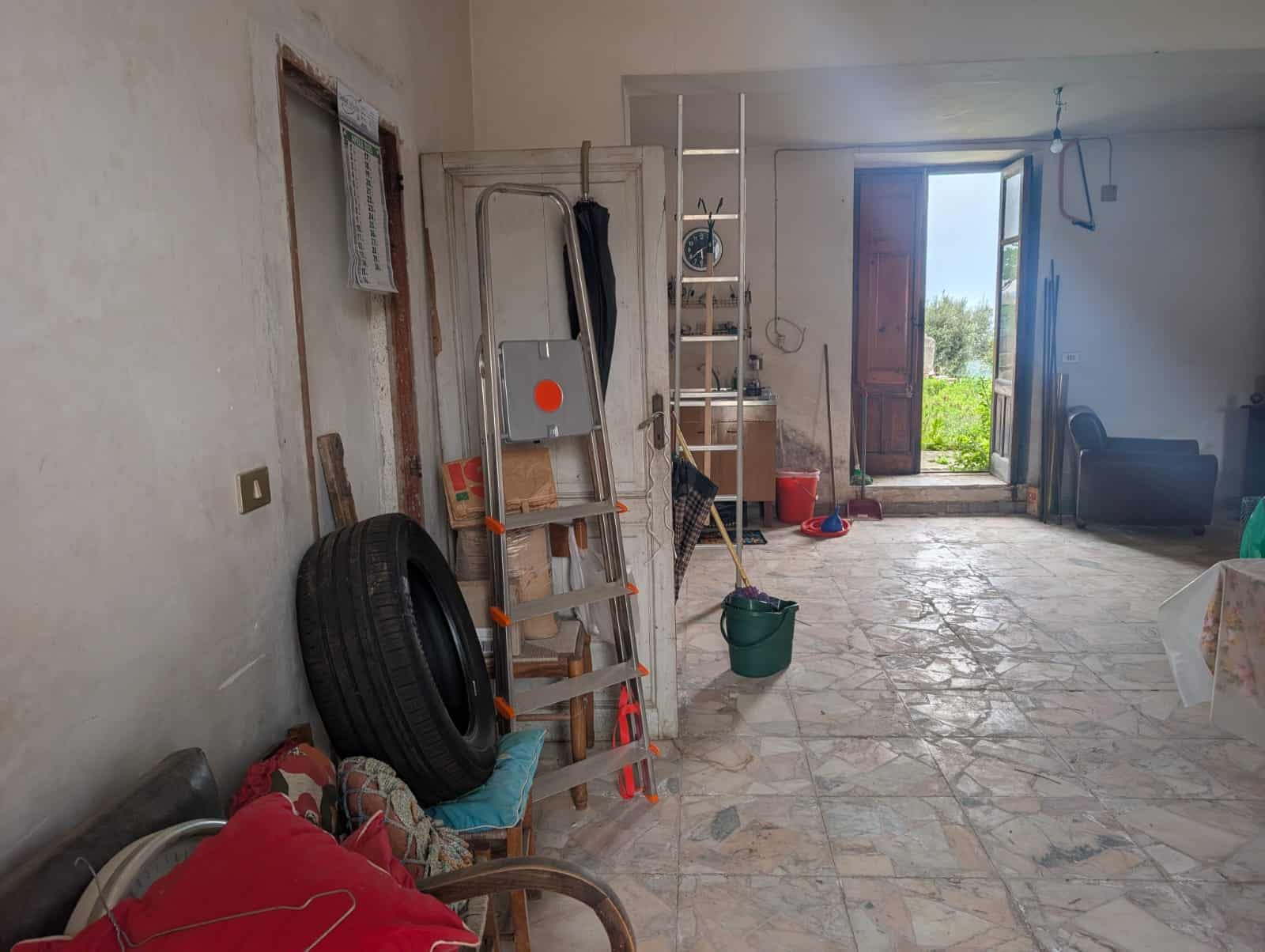 Interno di una casa in fase di ristrutturazione con attrezzi e materiali di lavoro, pareti non rifinite e pavimento in pietra, vista su un giardino attraverso una porta aperta.