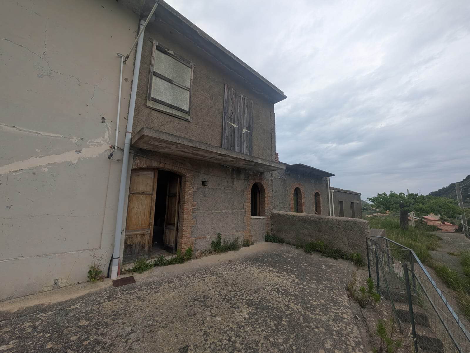 Vecchio edificio in pietra con finestre e porta in legno, situato in un'area rurale con vista su colline e cielo nuvoloso, ideale per ristrutturazione o investimento immobiliare.