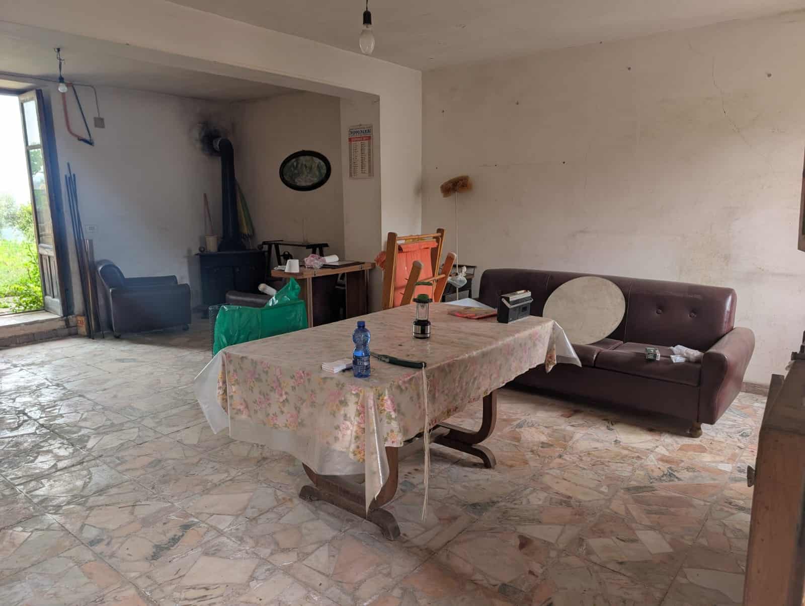 Interno di una casa con soggiorno semplice, tavolo con tovaglia floreale, divano e mobili di base, ambiente informale e poco arredato, luce naturale, pavimento in piastrelle.