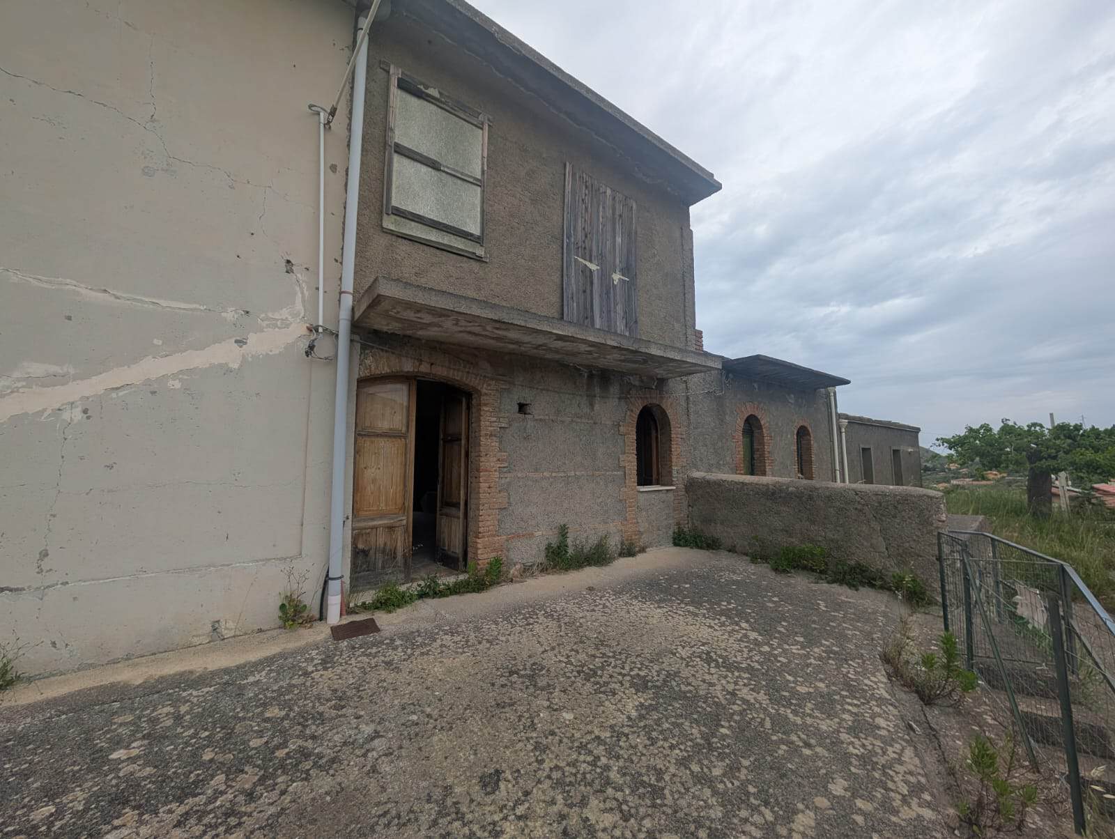 Vecchio edificio residenziale in pietra con facciata in parte in cemento, ingresso principale con porta in legno, finestre e balcone al primo piano, ambiente esterno con strada in pendenza e ringhiera.