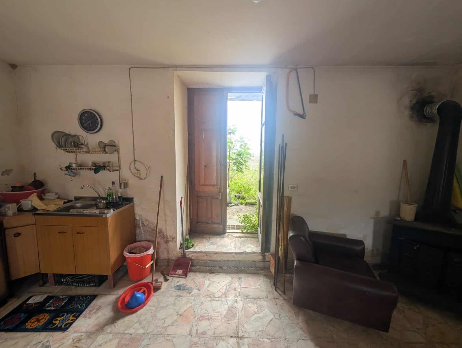 Interno di una stanza rustica con porta aperta che conduce a un giardino, arredamento semplice e pareti bianche, atmosfera casalinga e accogliente.