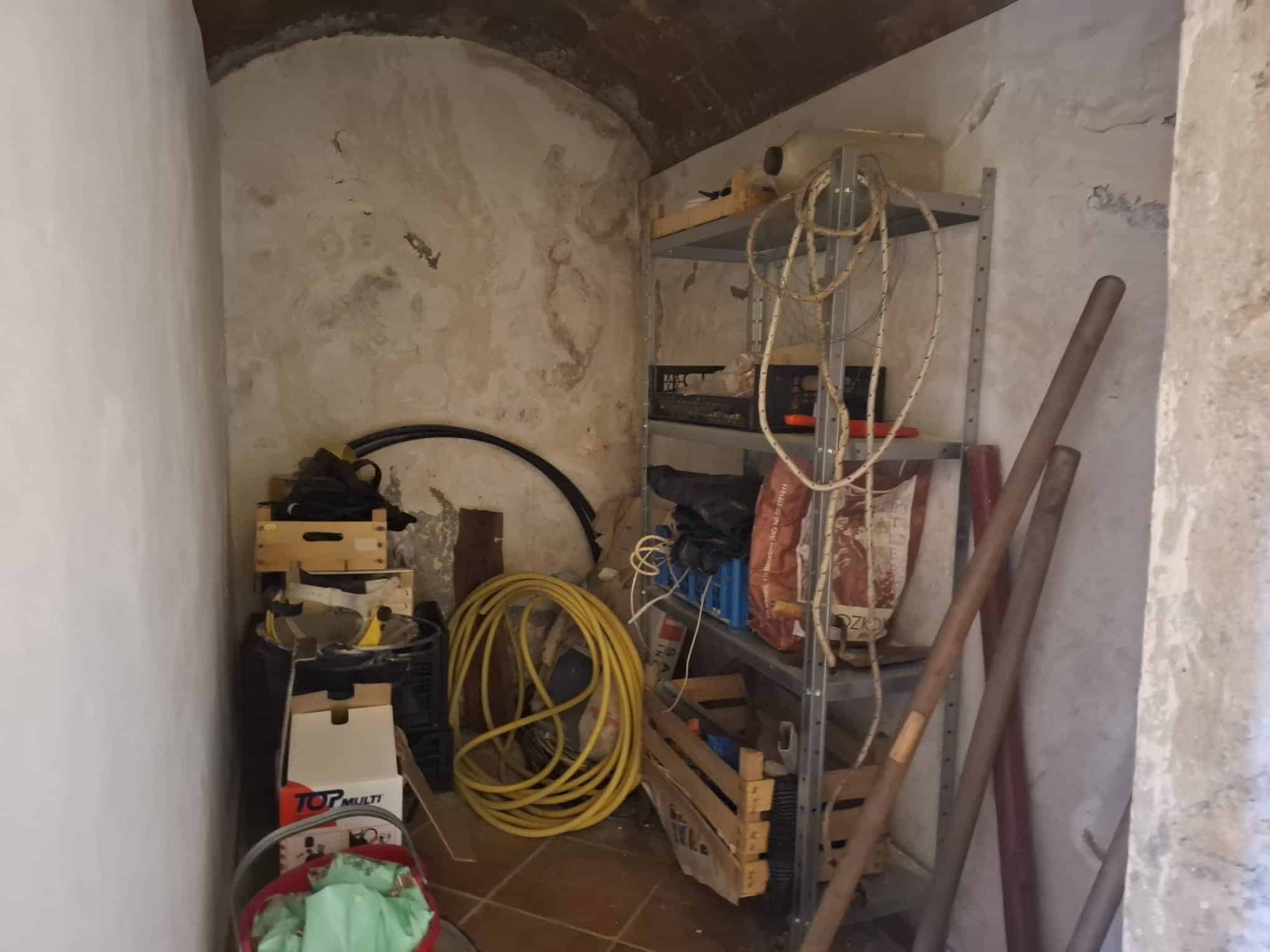 Riparazioni e attrezzi da lavoro in un magazzino o ripostiglio, con scaffalature piene di materiali, cavi e utensili, in un ambiente rustico con pareti in pietra e soffitto a volta.