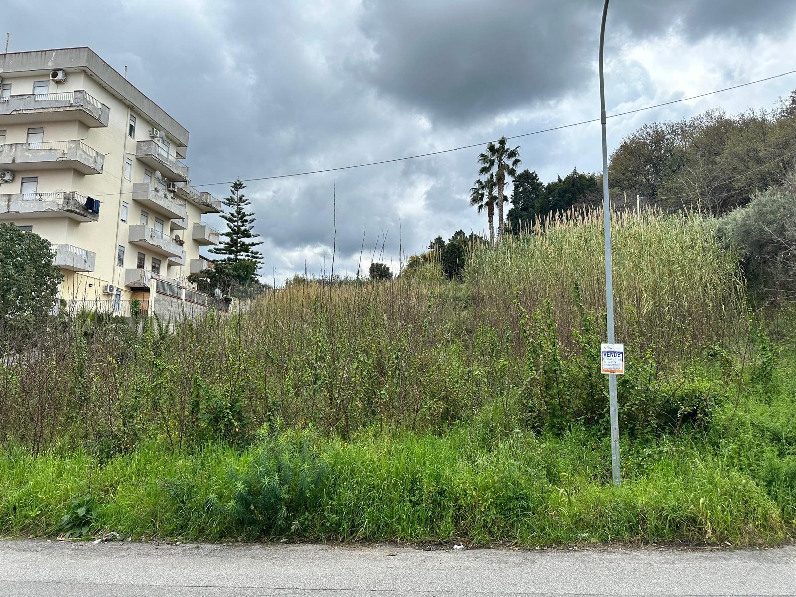 Terreno edificabile in vendita a Barcellona P.G. con vegetazione e lampione, vicino a edificio residenziale.