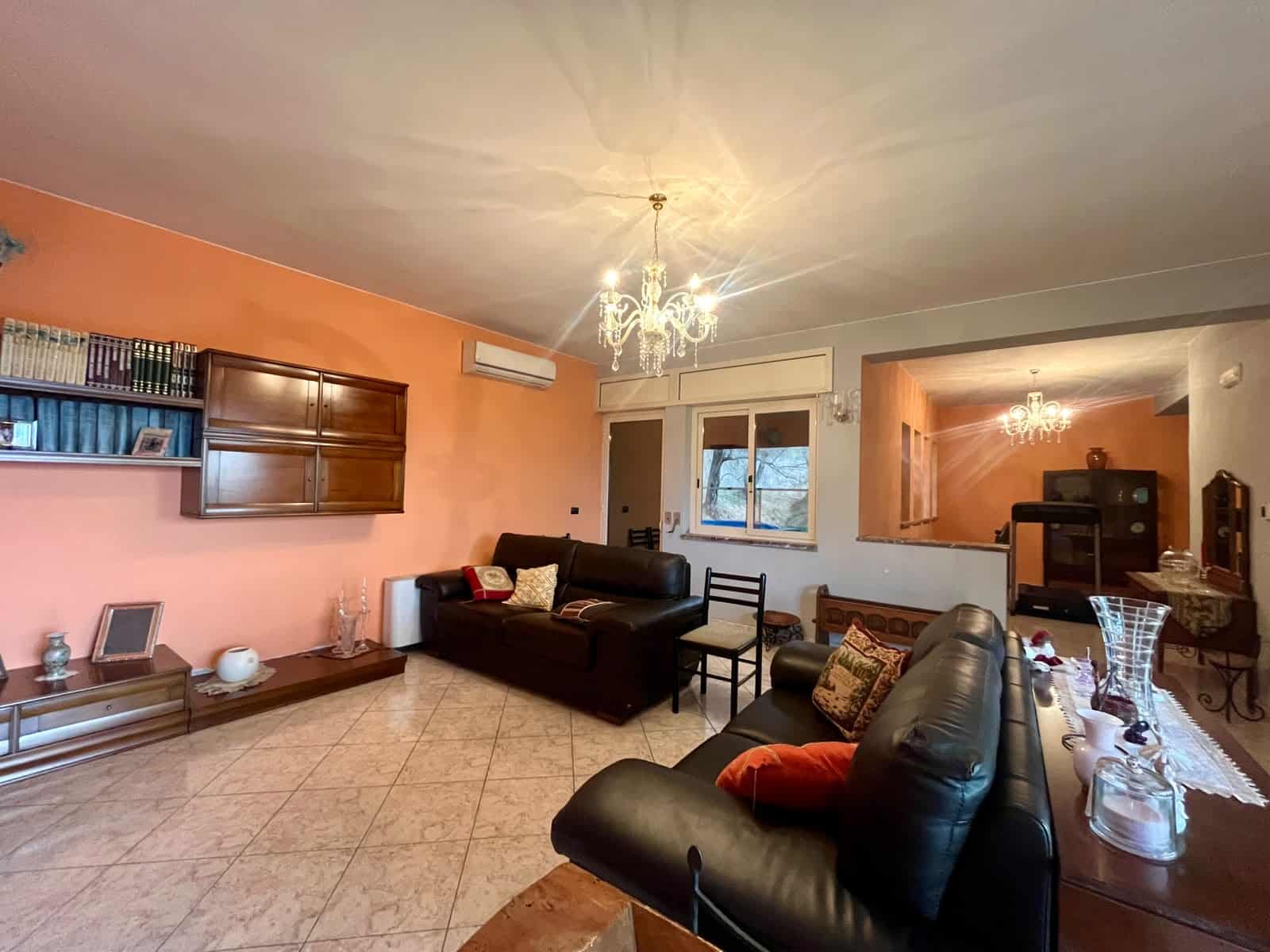 Elegante soggiorno con pareti color pesca, divani in pelle nera, libreria con libri e decorazioni, illuminazione a chandelier, e ampie finestre che lasciano entrare luce naturale.
