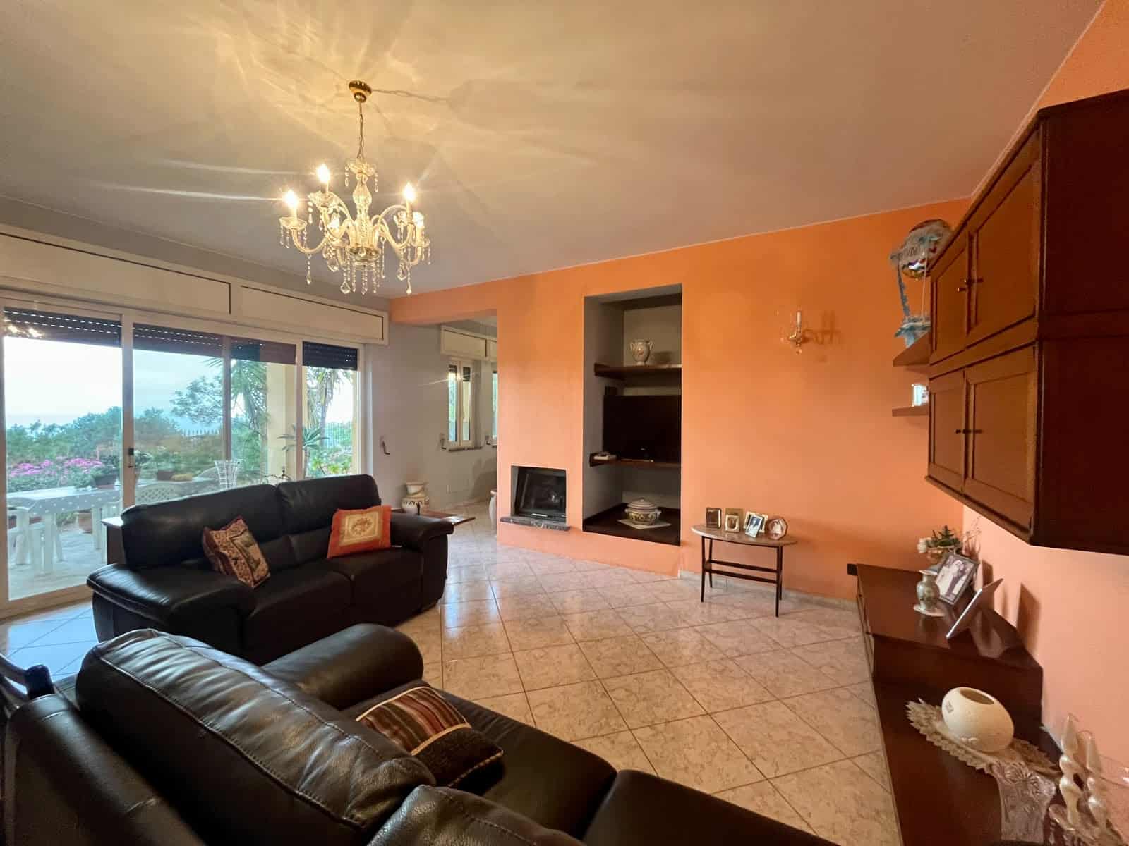 Elegante soggiorno con pareti arancioni, divani in pelle nera, camino e mobili in legno, illuminato da lampadario a chandelier, con grandi finestre che offrono vista sul giardino e luce naturale.