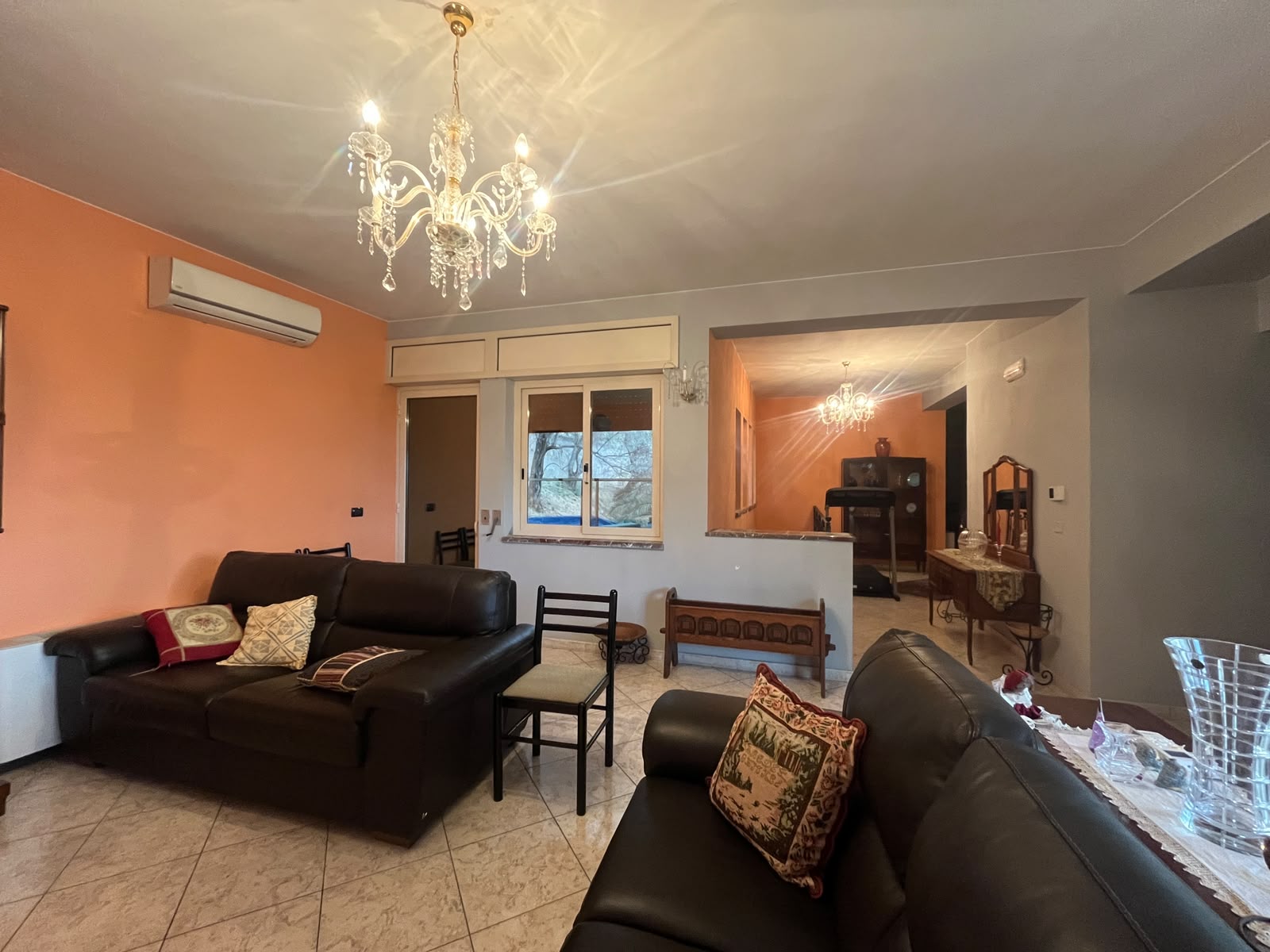 Interno soggiorno con divani in pelle nera, lampadario elegante e pareti color pesca, con finestre che offrono luce naturale e vista sul giardino.