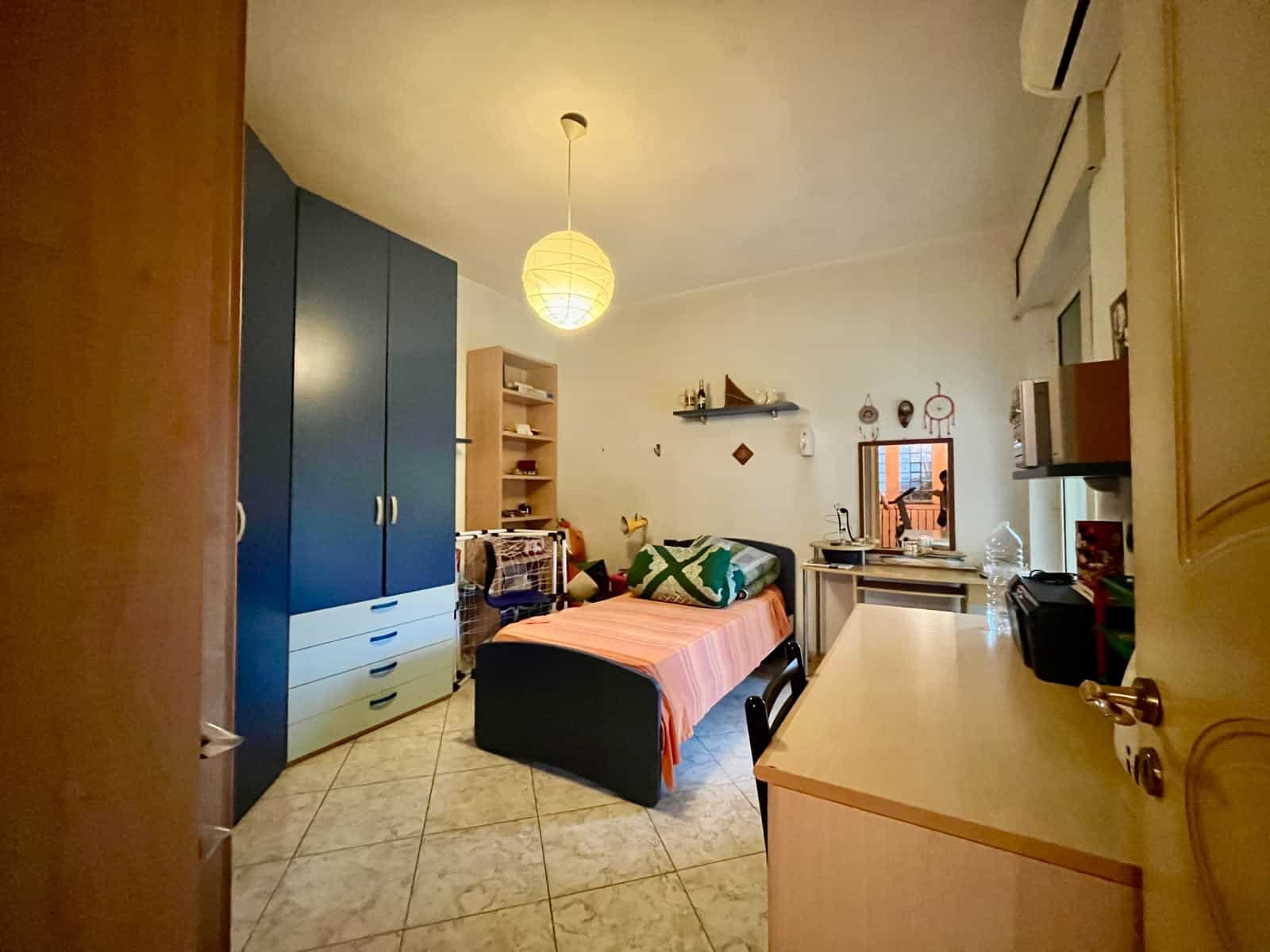 Una stanza da letto con arredi moderni, armadio blu, letto con copriletto rosa, scrivania e finestra che lascia entrare luce naturale, in un appartamento in stile contemporaneo.